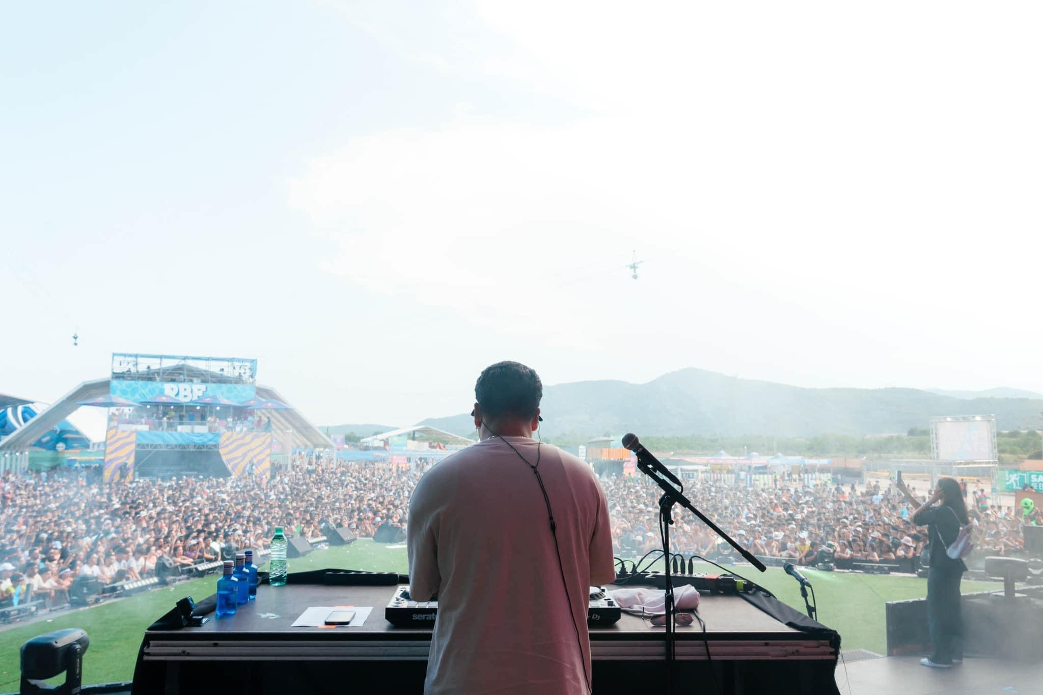 Person DJing at an outdoor event with a large crowd and stage in the background, facing away from the camera.