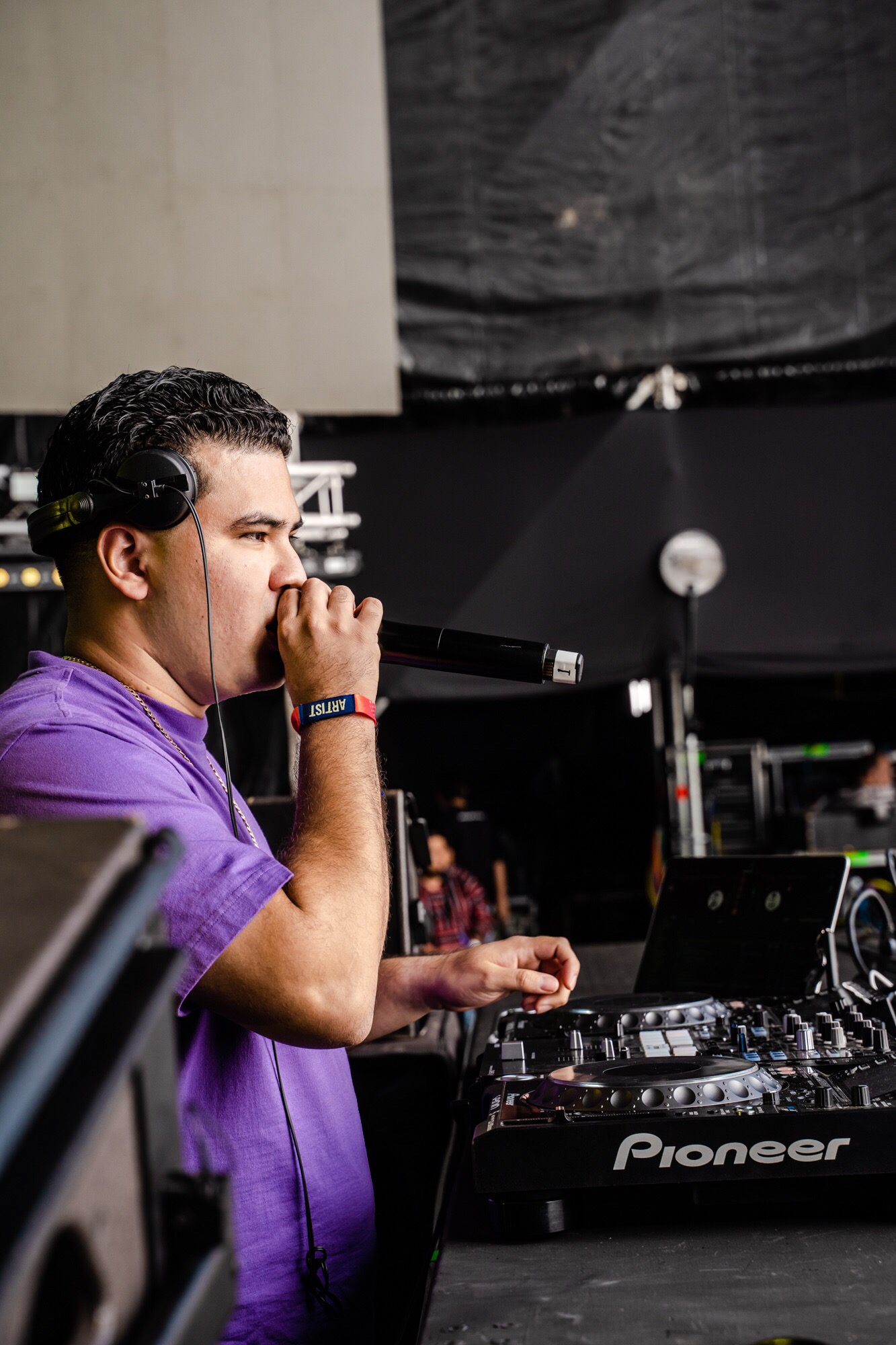 Young man with headphones holding microphone, standing at DJ equipment in a dark room, possibly a music studio or club.