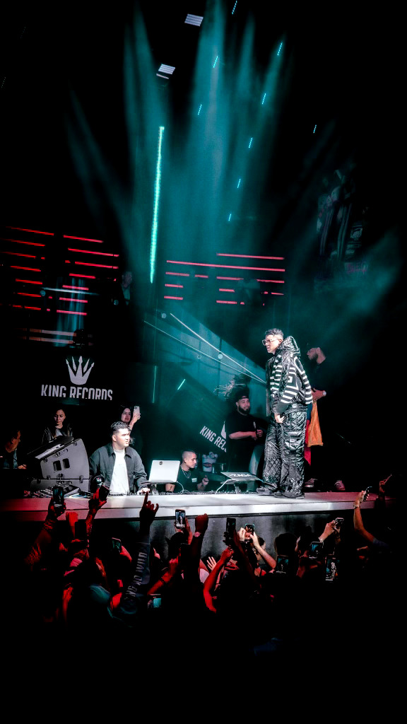 Performer on stage with bright blue and white lights, audience in foreground, and a backdrop with red lines.
