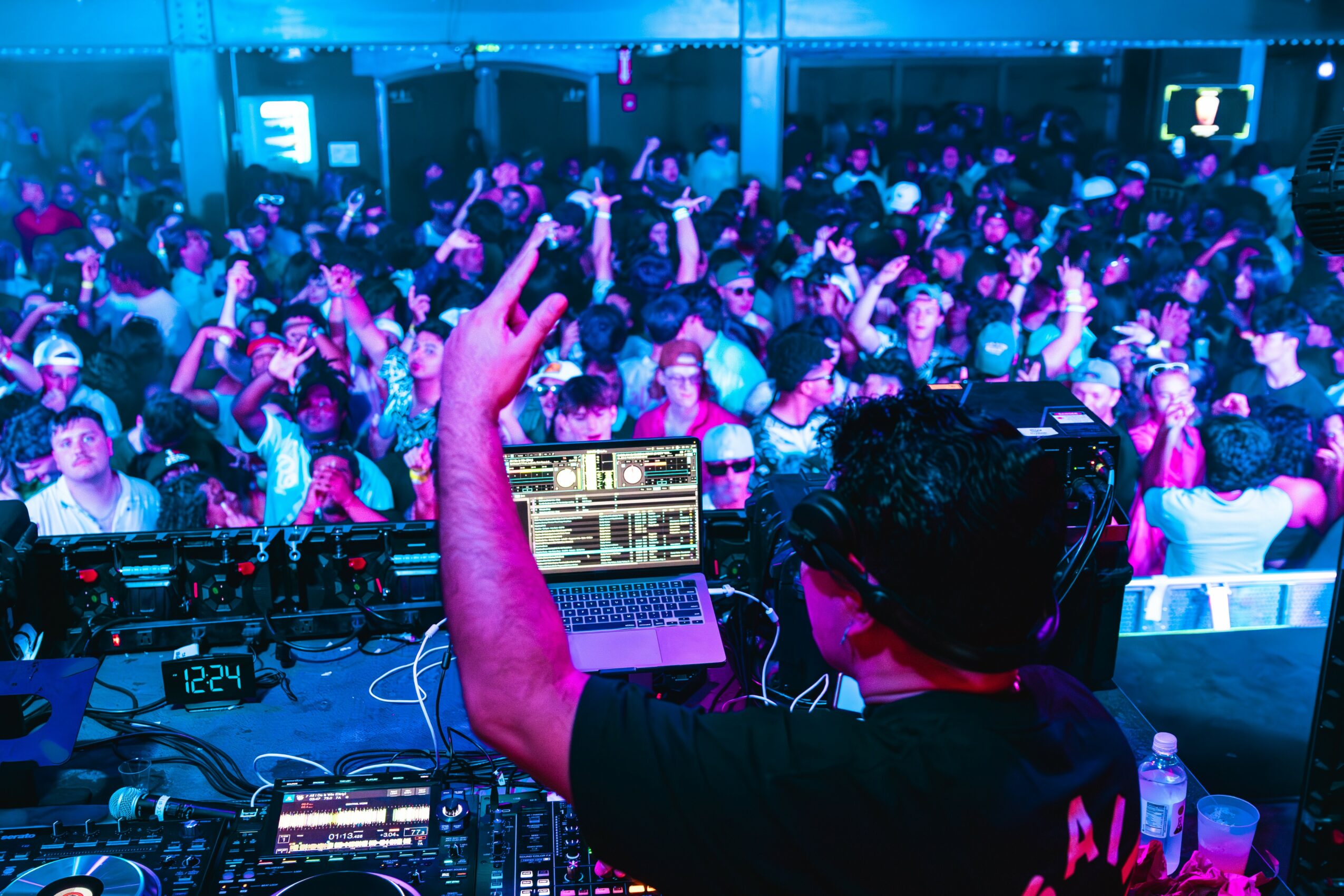 Crowd of people at a concert or event, with a DJ or performer in the foreground, raising a hand.
