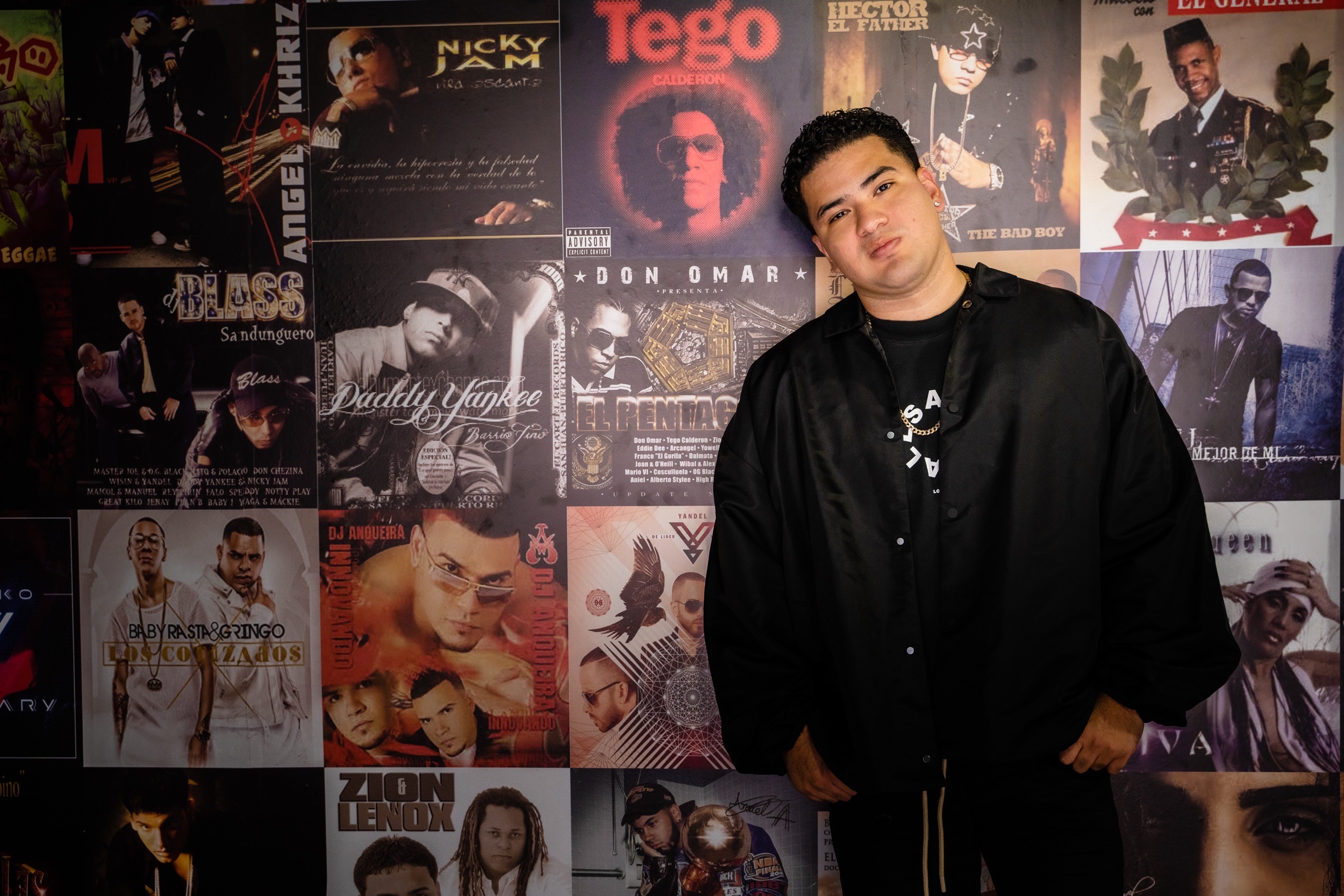 Young man in black jacket standing in front of a wall covered with various music album posters.