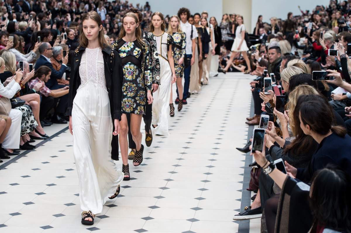 Models walk down runway during fashion show, audience seated on both sides taking photos, large crowd present.