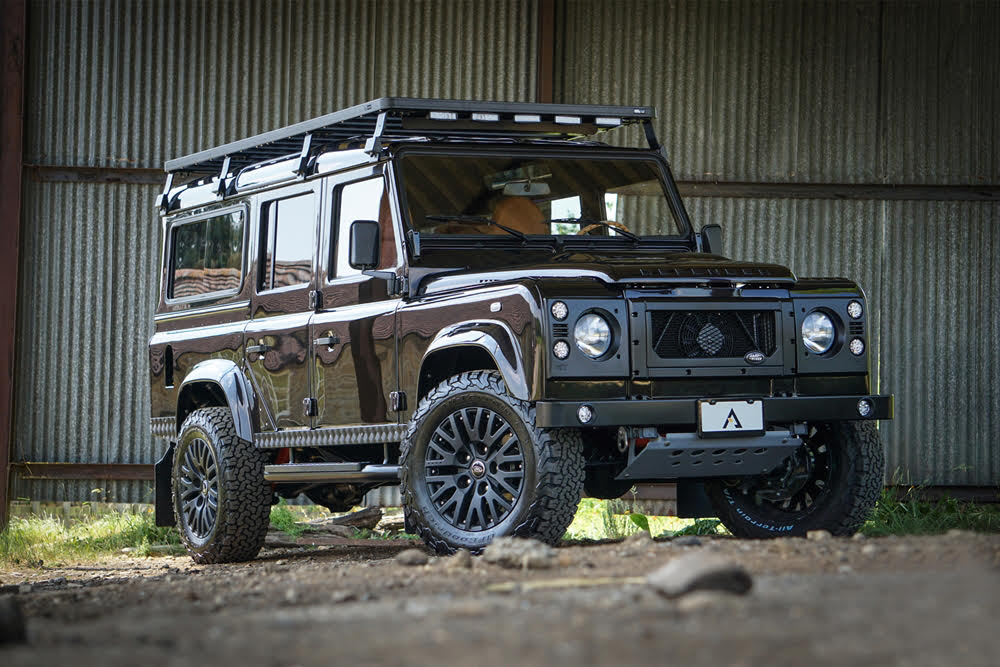 Black off-road vehicle parked on ground in front of a corrugated metal wall, with a roof rack and large tires.