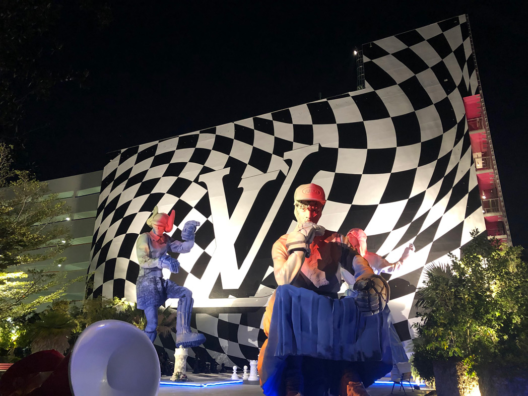 Nighttime scene with people in front of a large checkered building and a prominent logo, surrounded by trees and lights.