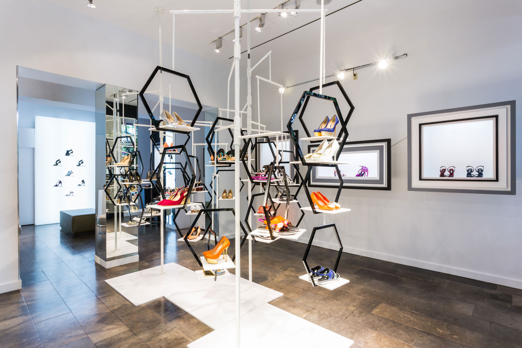 Display of shoes on black hexagonal shelves in a gallery with framed artwork on white walls and dark flooring.