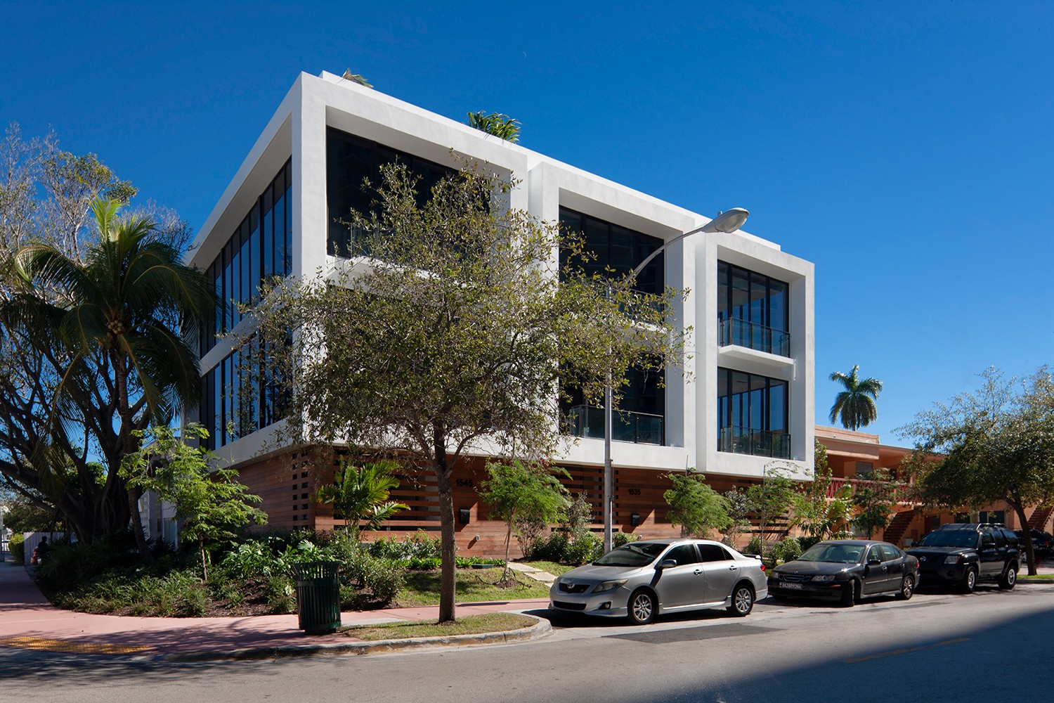 Modern multi-story building with large windows, trees, and parked cars in front, under a clear blue sky.