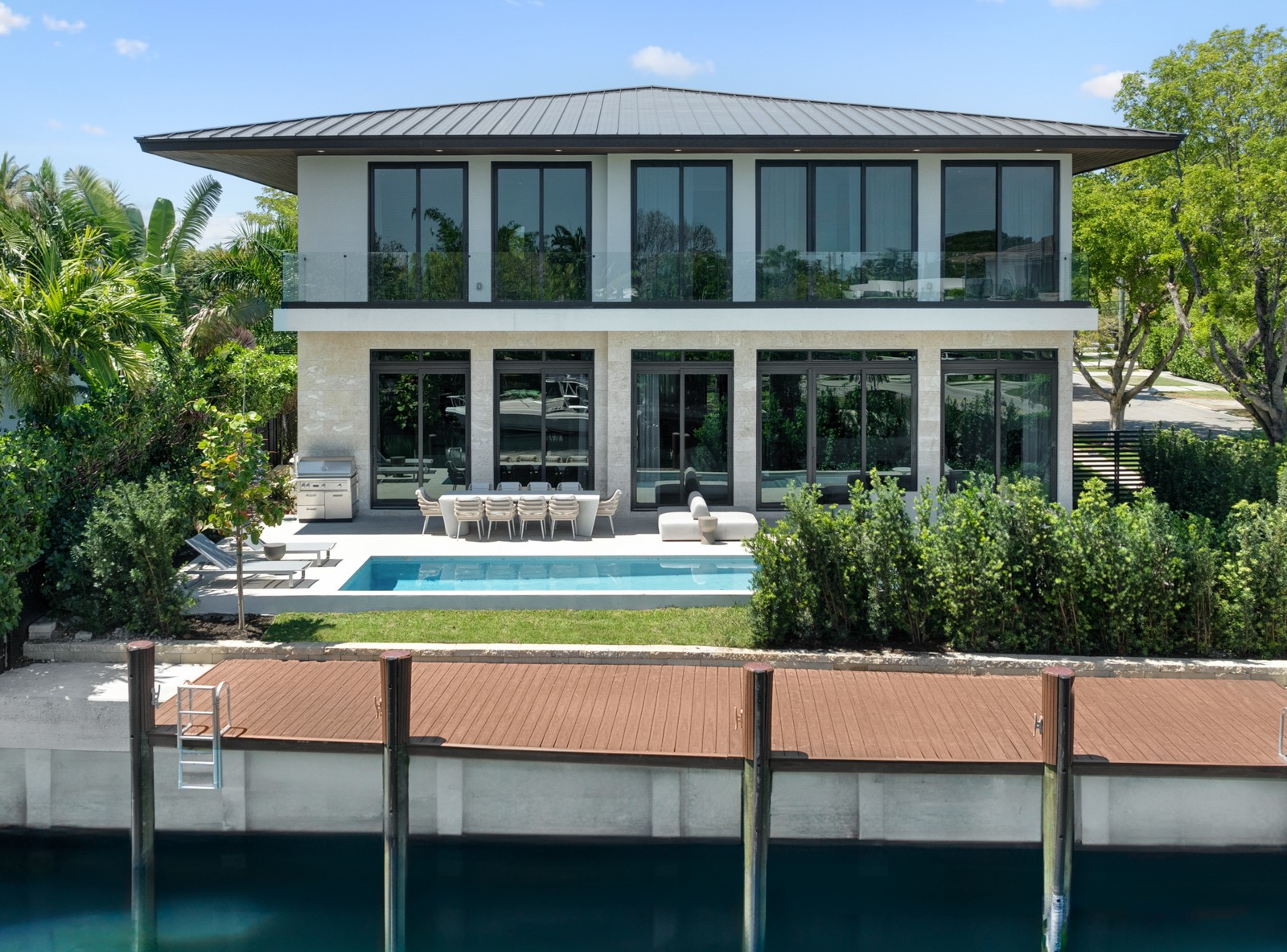 Modern two-story house with large glass windows, balcony, outdoor seating, and a swimming pool, surrounded by greenery.