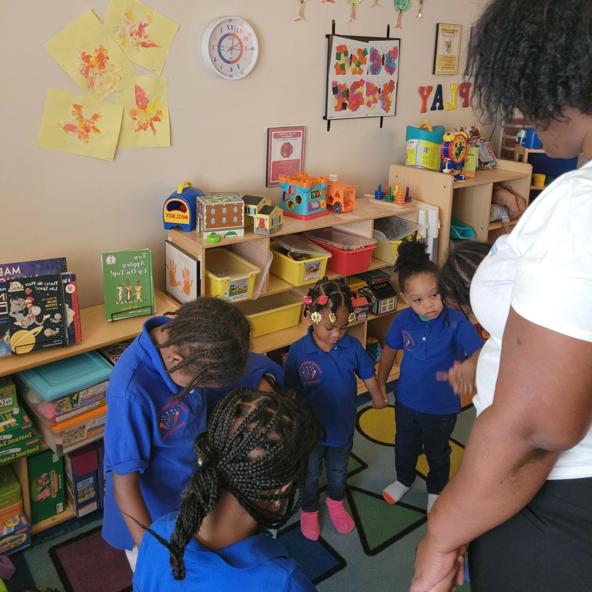 Four young children in blue uniforms stand in a classroom with colorful shelves and artwork on the wall.