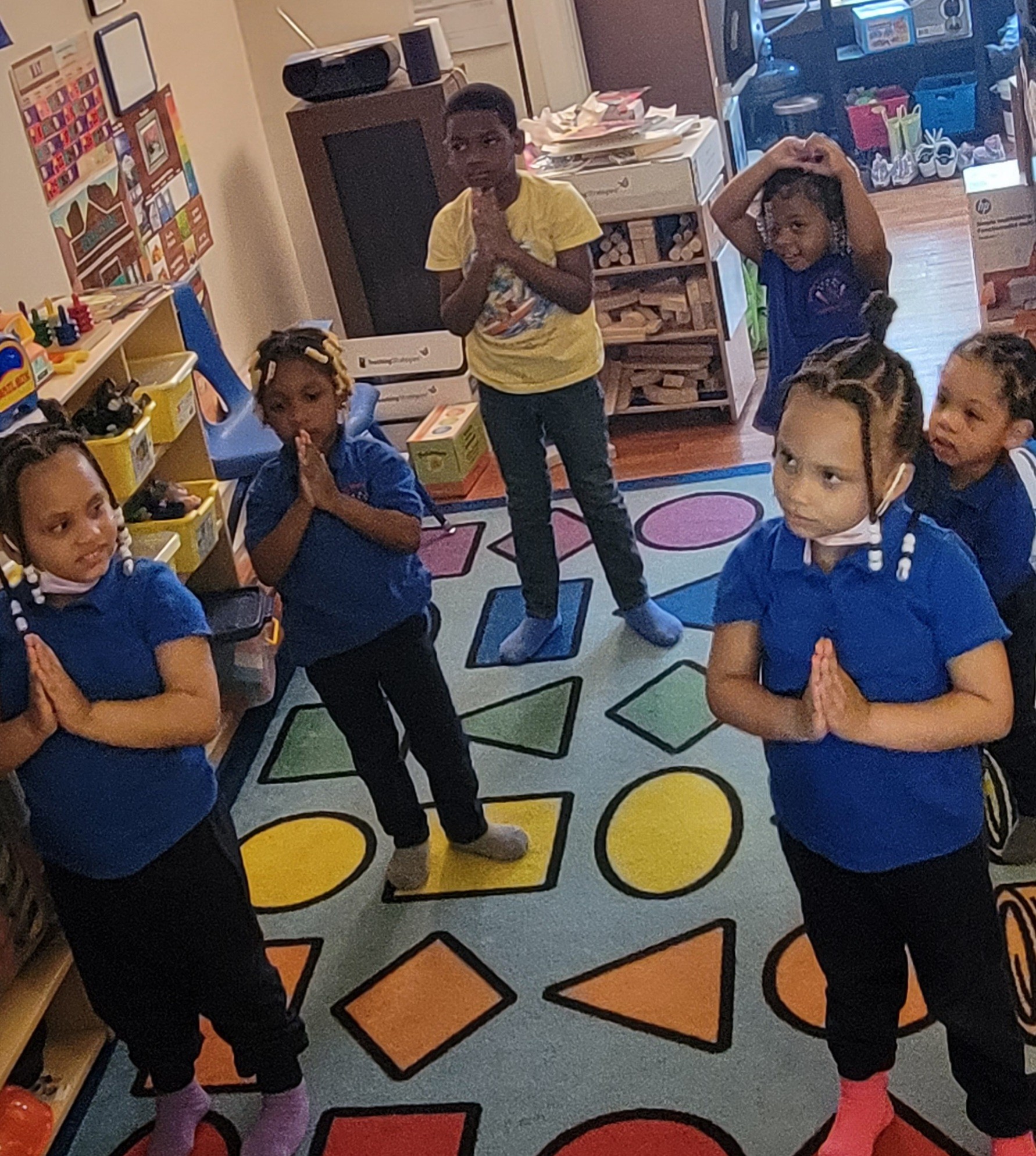 Group of children standing with hands together in a classroom, colorful rug and shelves visible in background.