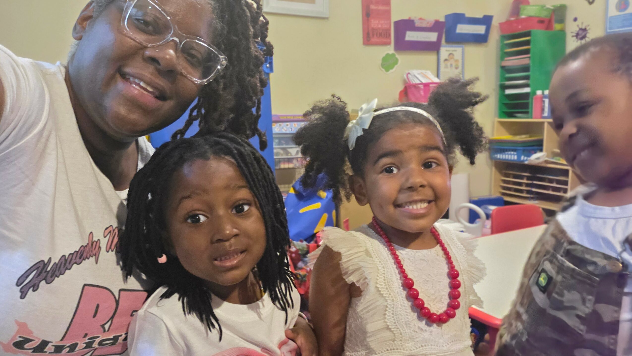 Four children and an adult smiling in a colorful classroom, with shelves and educational materials in the background.