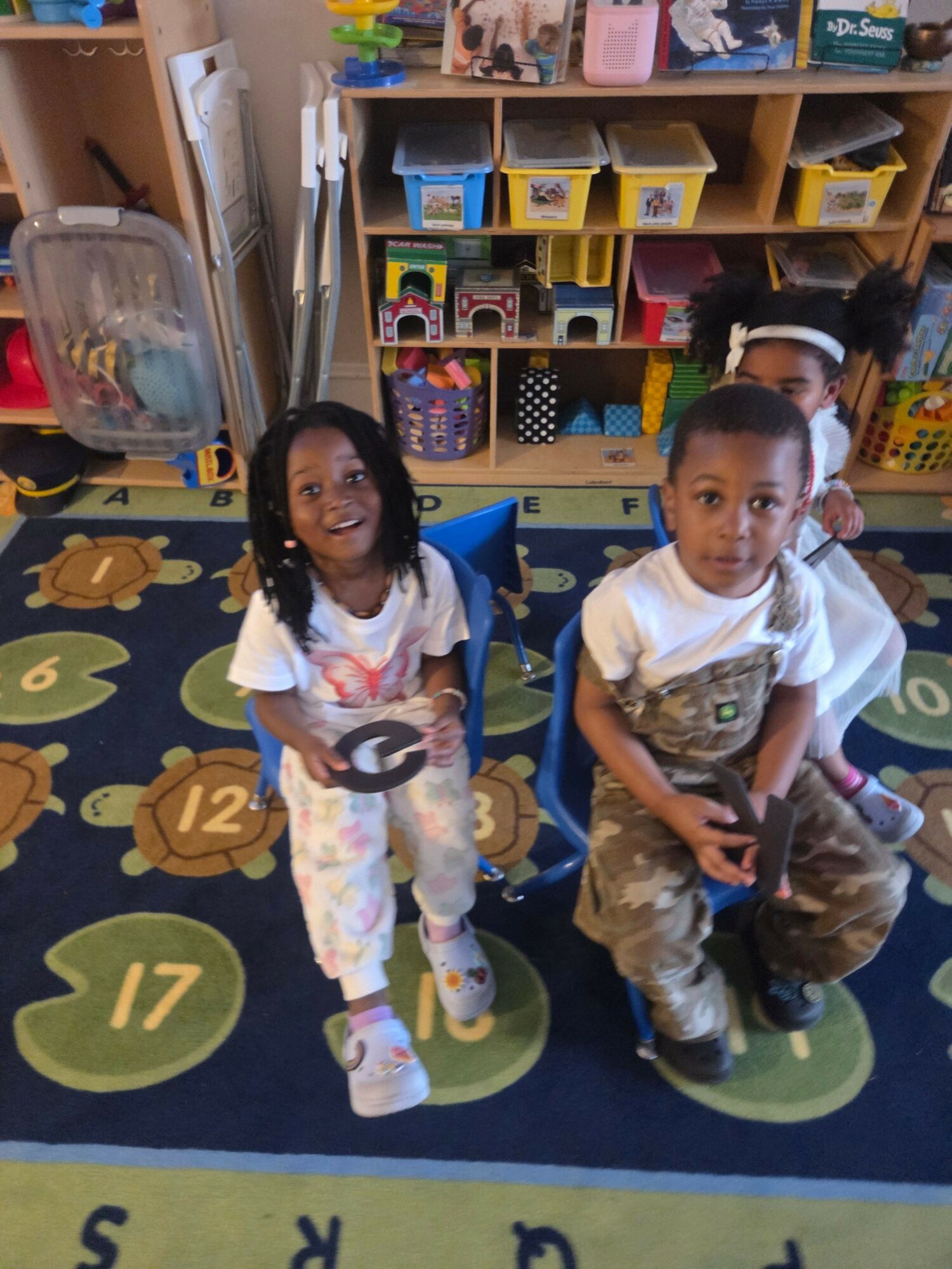 Two children in a classroom, one holding a toy gun, standing on a colorful circular rug with numbers and animals.