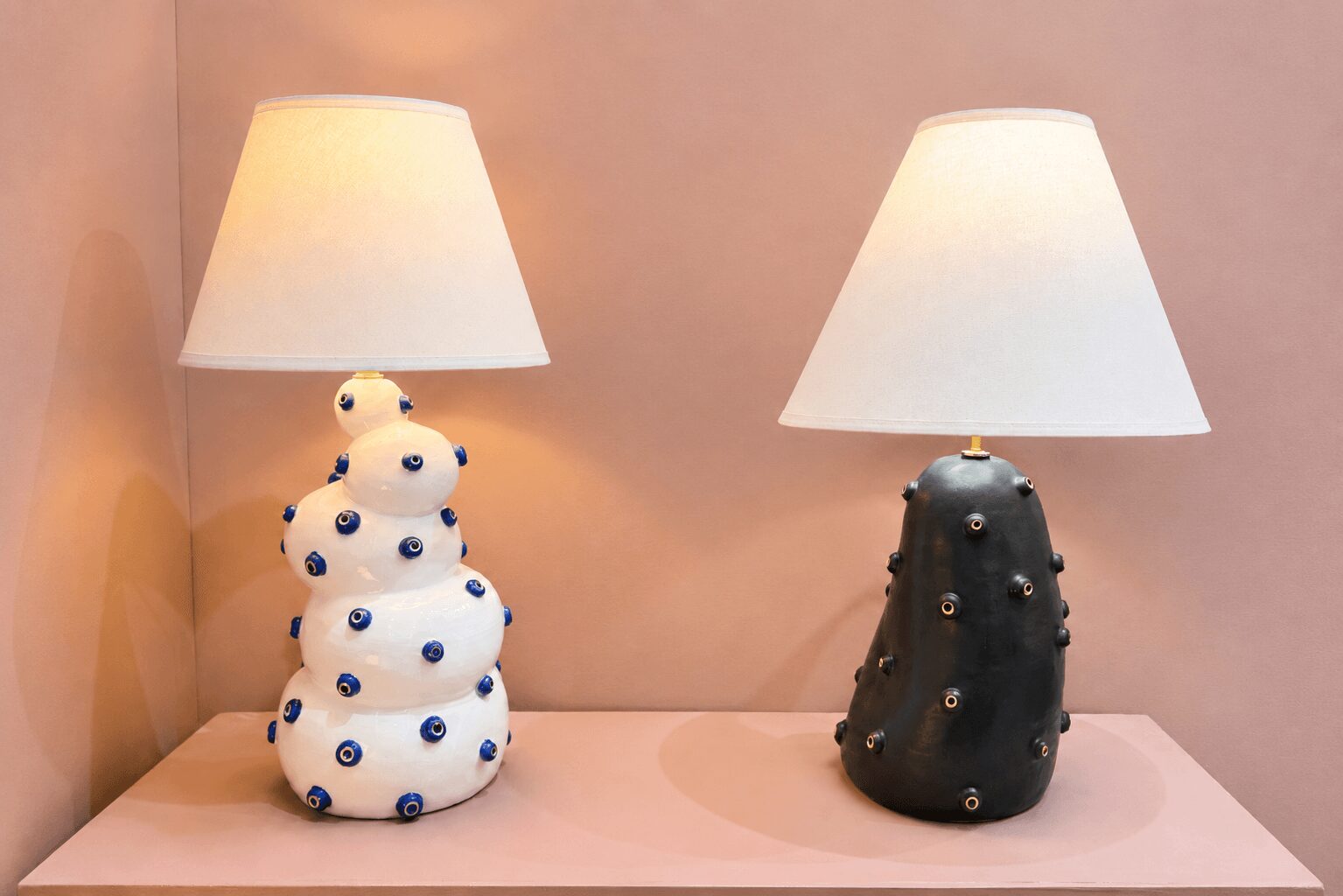Two table lamps with white shades and decorative bases, one white with blue accents, the other black with silver accents, on a pink surface.