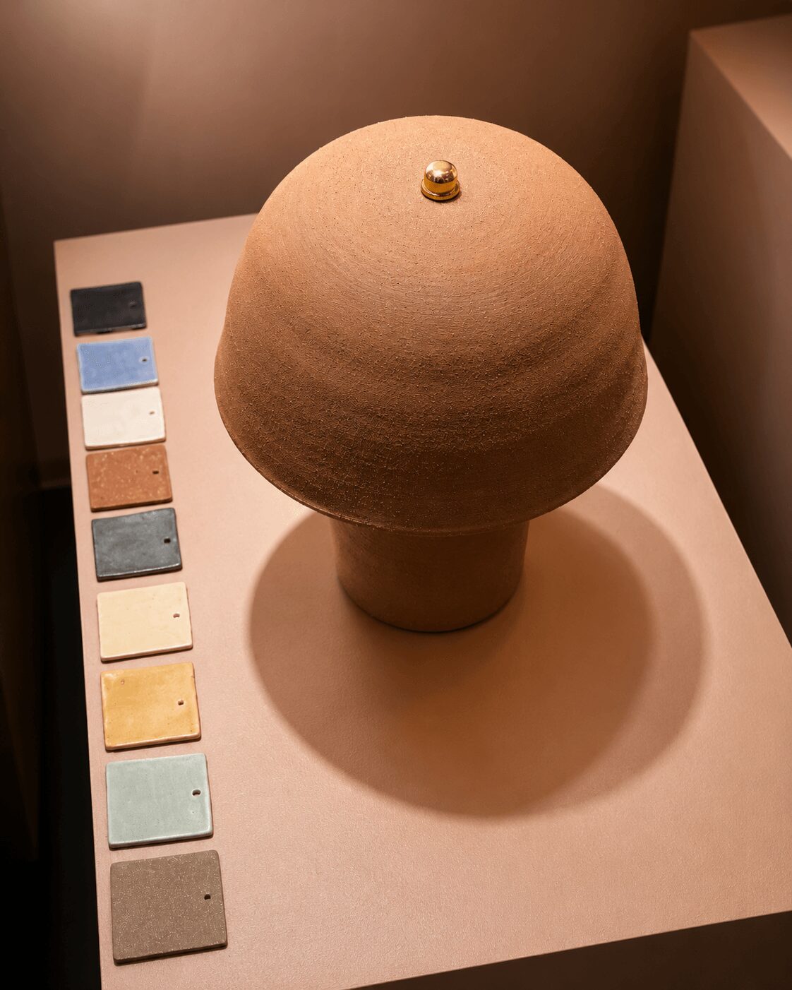 Round wooden lamp with a small gold knob on top, placed on a white surface with color swatches beside it.