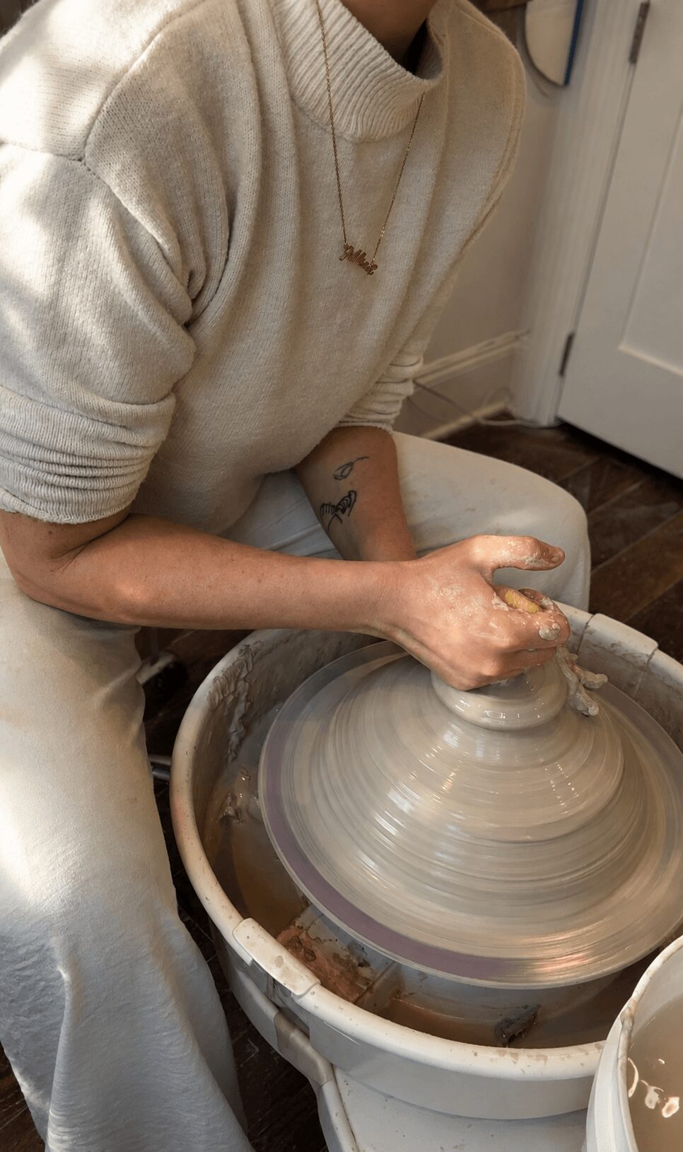 Person shaping clay on a pottery wheel, wearing a beige sweater, with a tattoo on their arm.