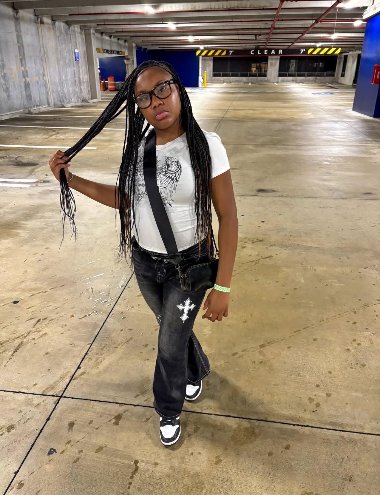 Young woman with long braided hair, glasses, white t-shirt, black pants, and sneakers, standing in an empty parking garage.