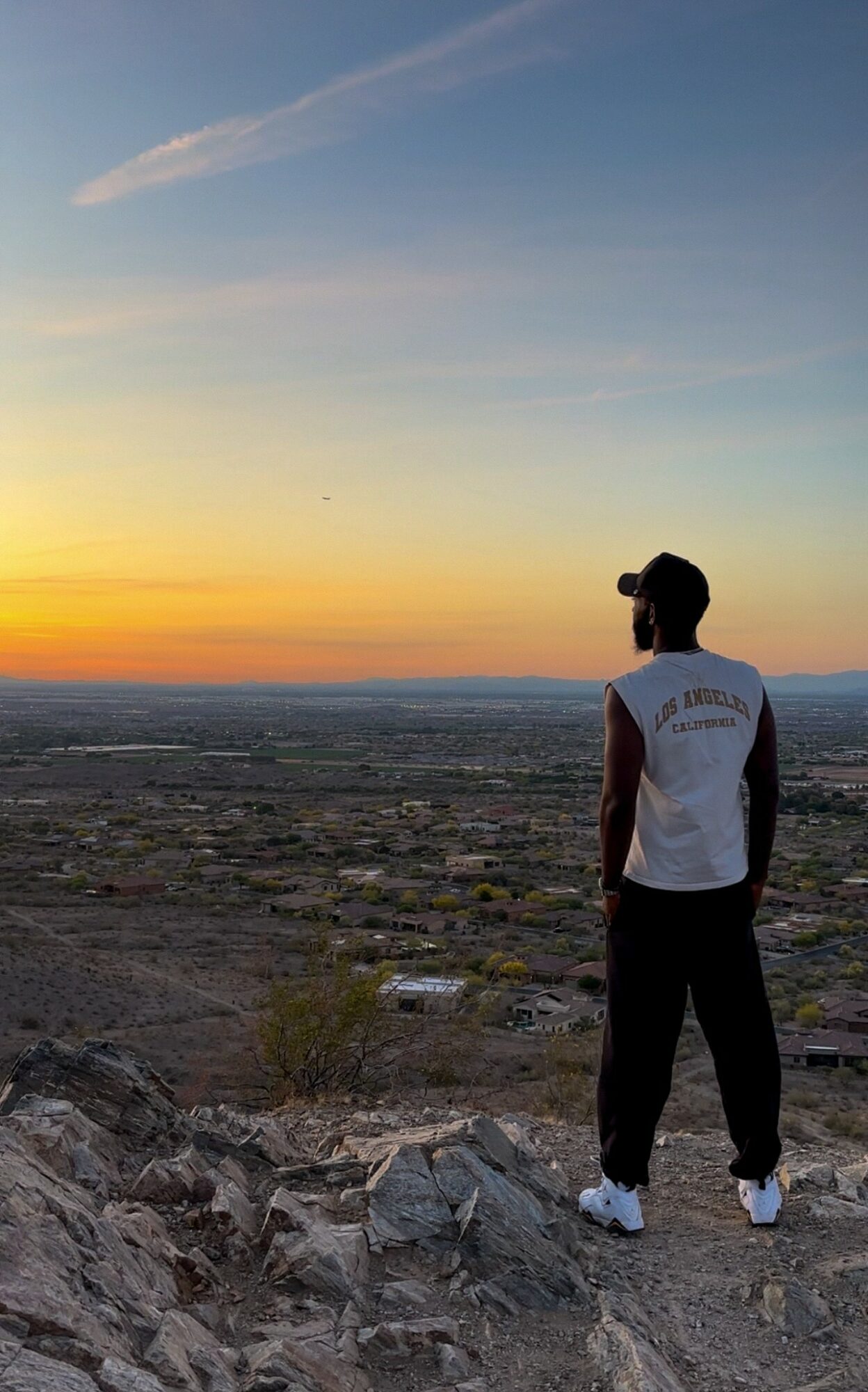 Person standing on rocky terrain watching sunset over cityscape, wearing a sleeveless shirt and cap, with a colorful sky.