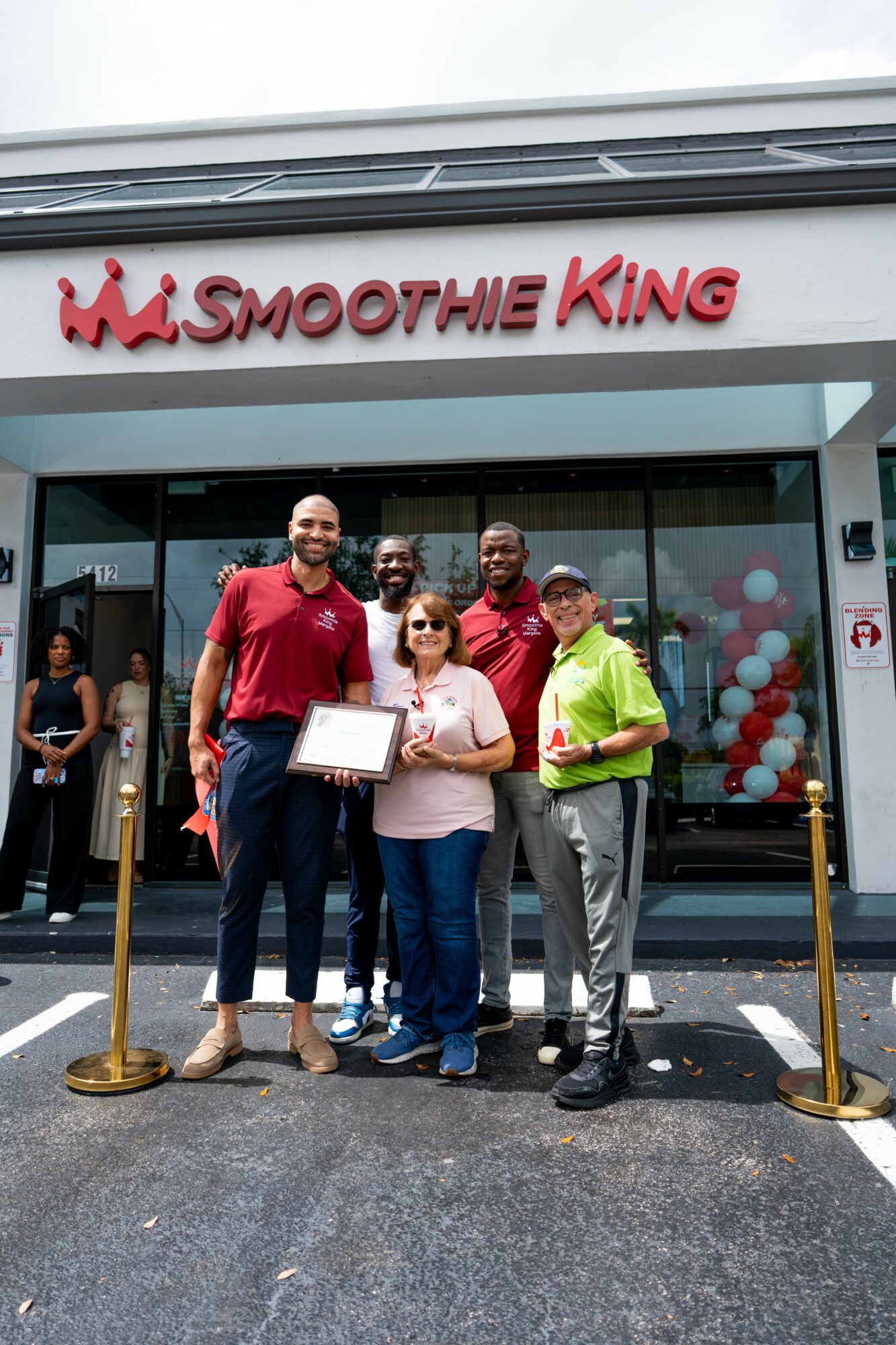 Group of six people standing outside Smoothie King store, holding a certificate, smiling, with balloons inside the store visible through glass.