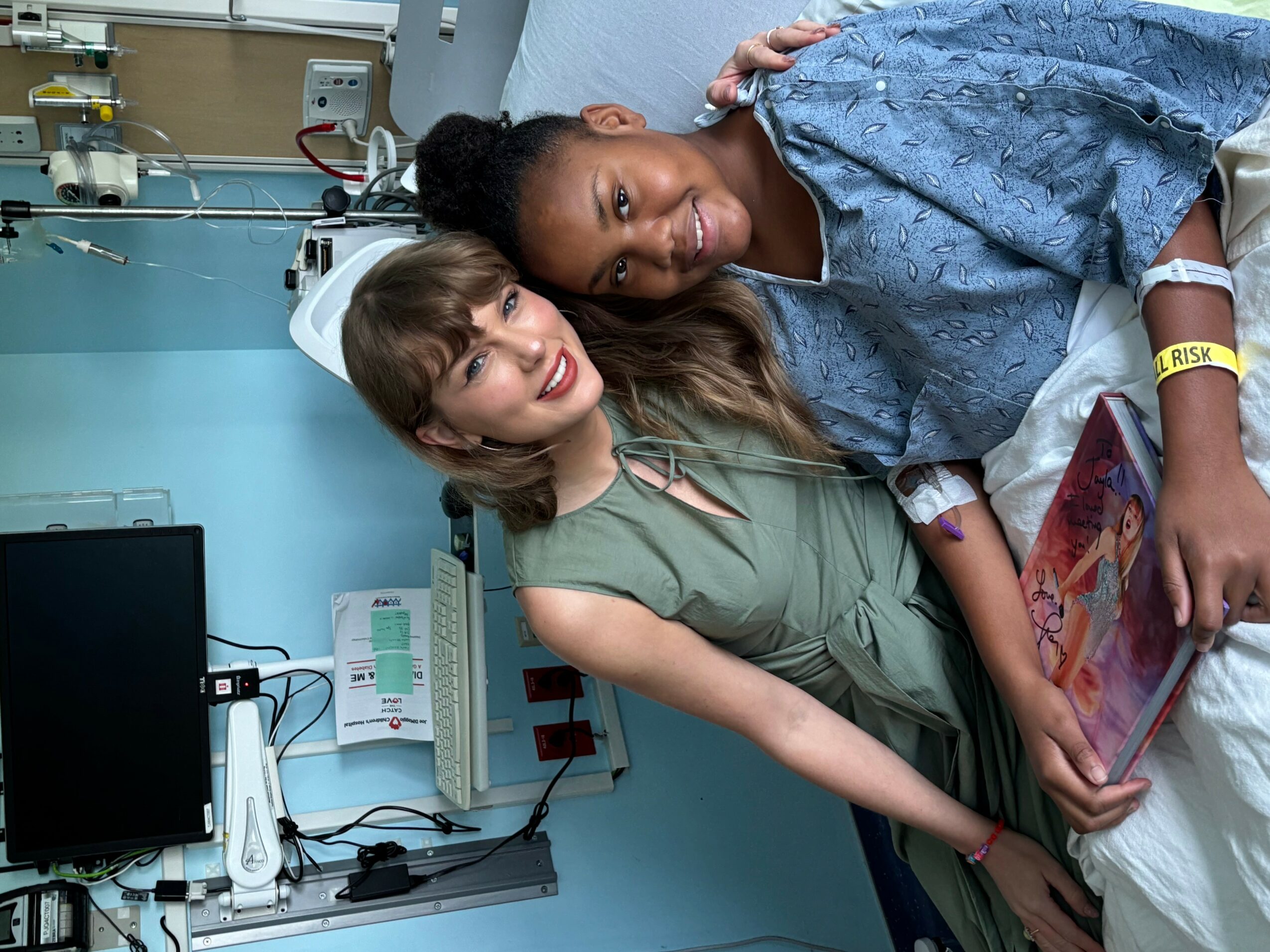 Two women in a hospital room, one in a hospital gown, the other in casual clothing, smiling and leaning together.