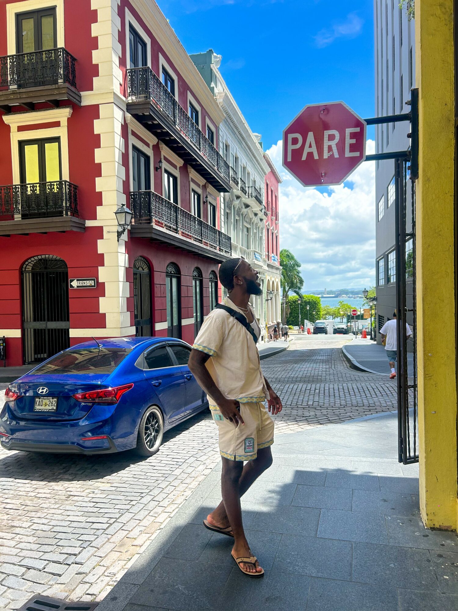 Person standing on sidewalk near a stop sign, colorful buildings in background, blue sky, parked car nearby.