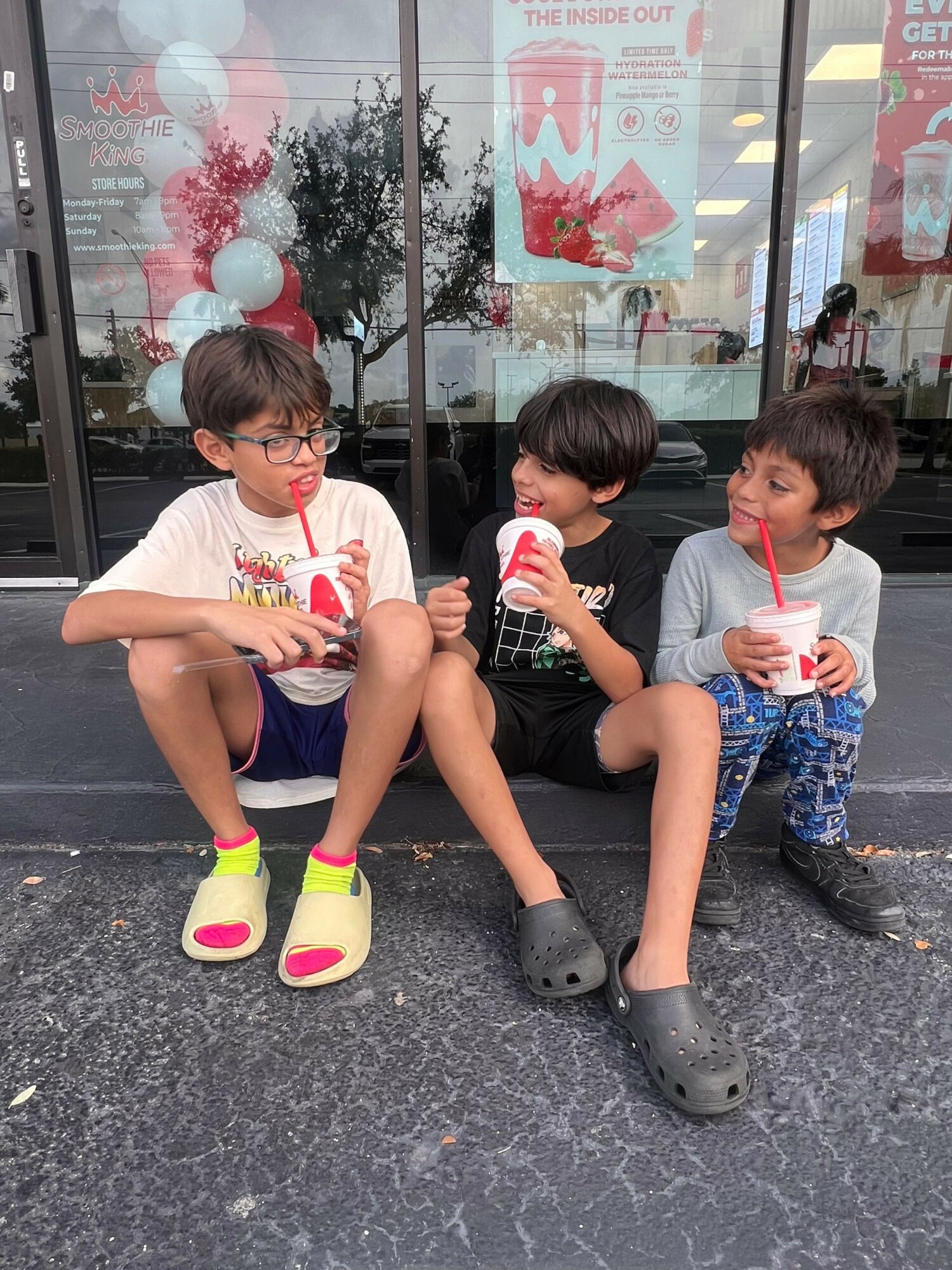 Three children sitting on the sidewalk, drinking from bottles, with a store window and balloons in the background.