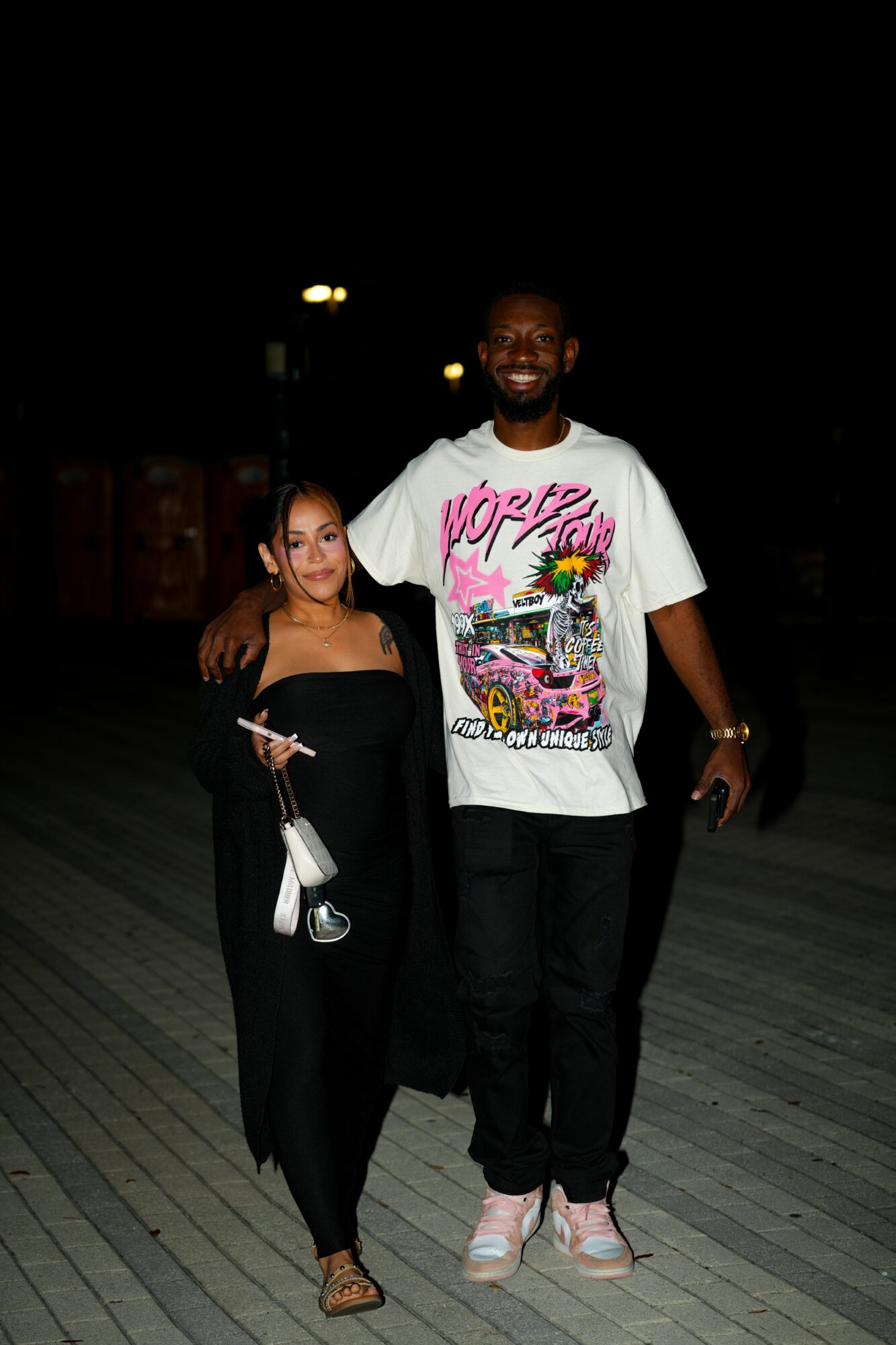 A man and woman standing outdoors at night, smiling, with streetlights in the background.