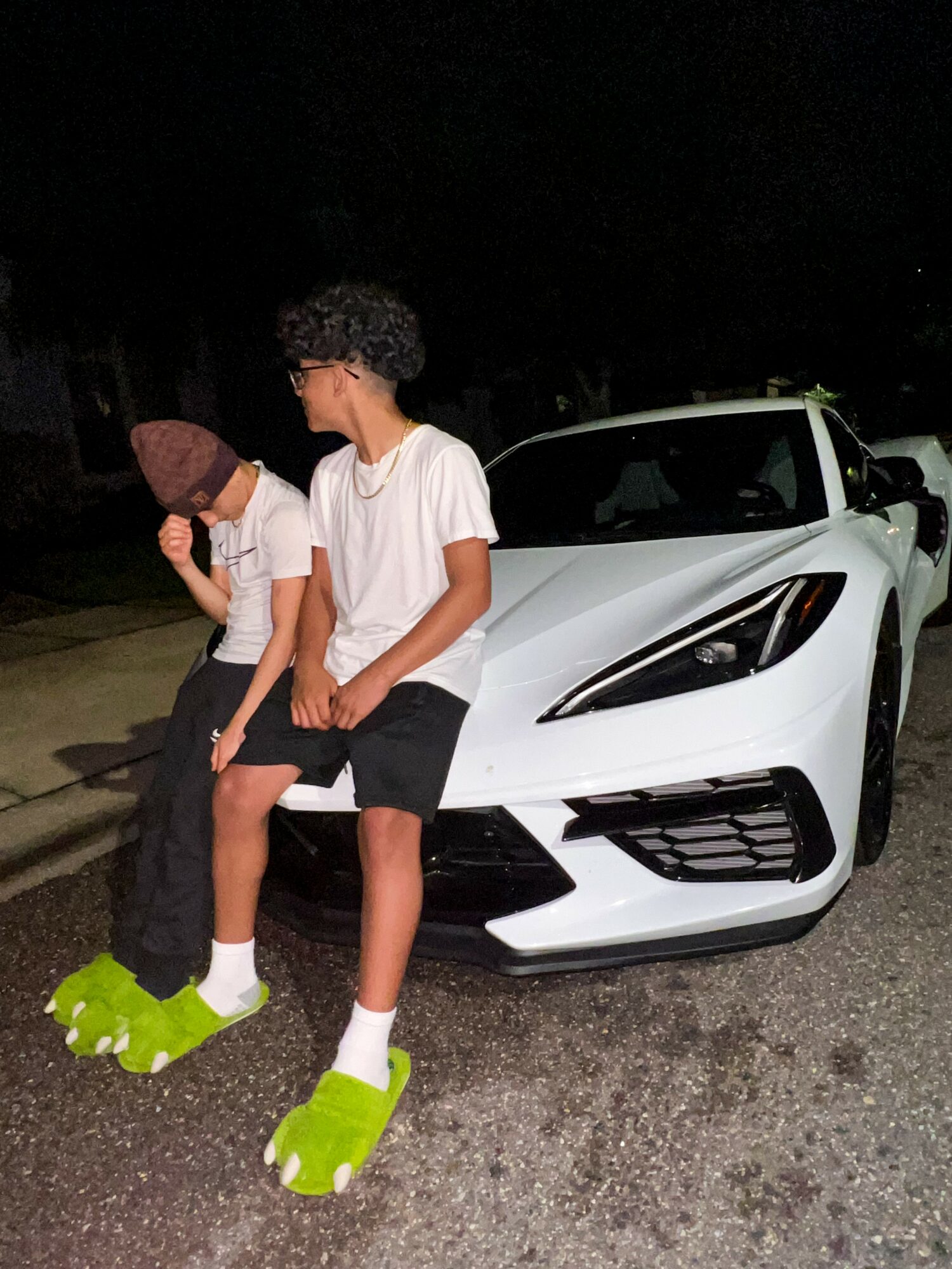 Two children sit on the front of a white sports car at night, one wearing green shoes and the other with brown shoes.