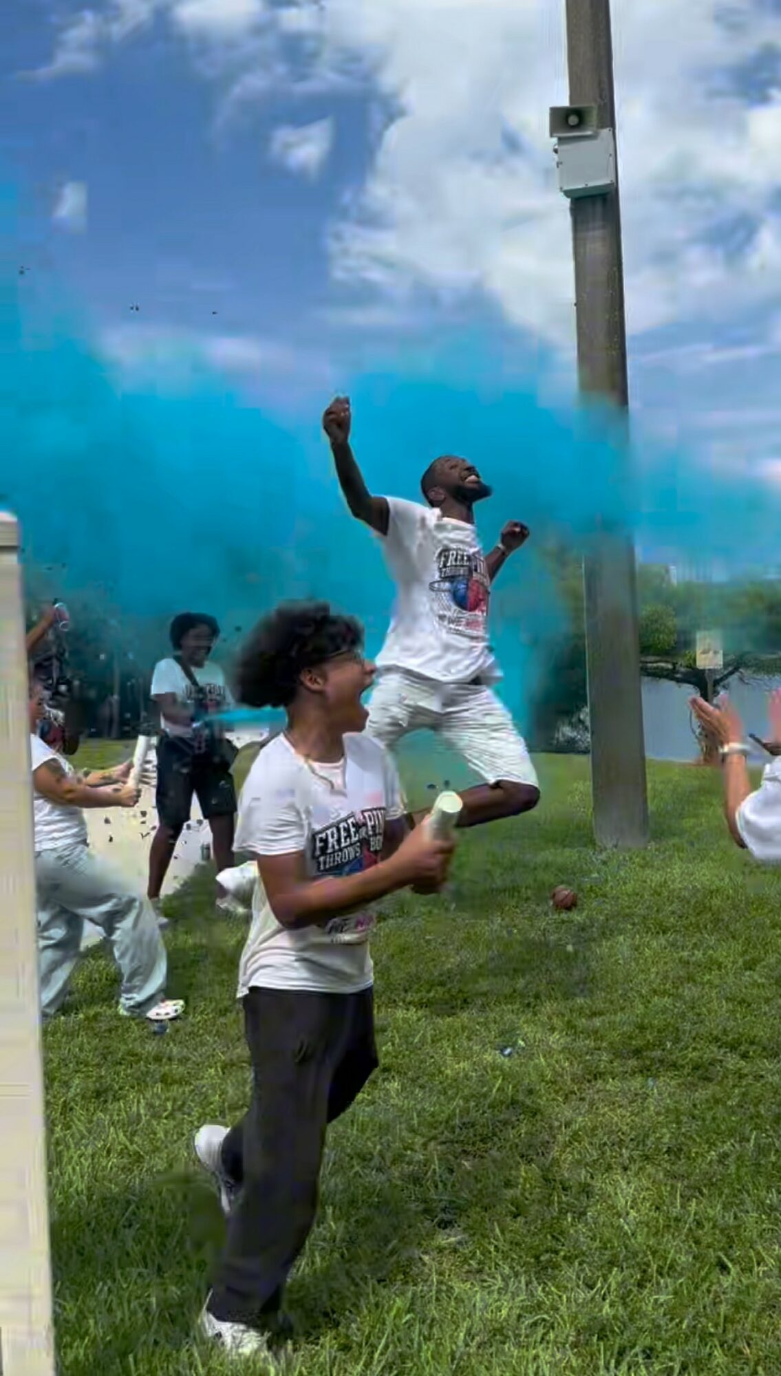 People celebrating outdoors with colored powder, one person jumping, others holding containers, grassy area, cloudy sky.
