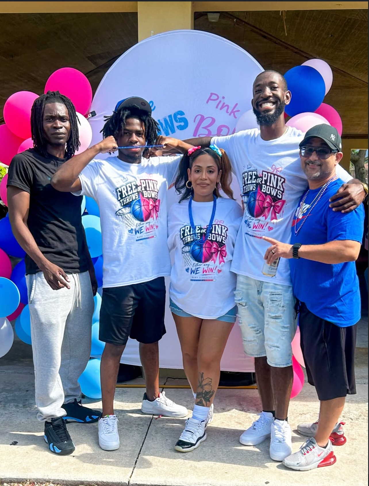 Group of five people standing outdoors in front of a pink, blue, and white balloon display, smiling and posing for the photo.
