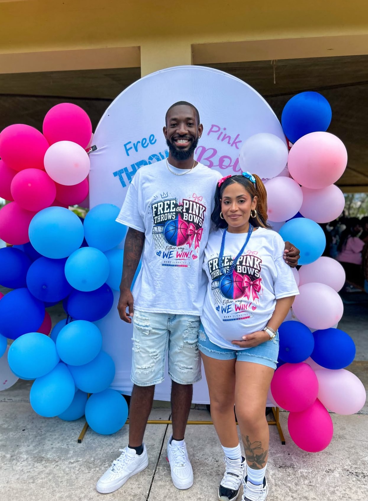 Two people, a man and a woman, stand in front of a balloon display with pink, blue, and white balloons, smiling.