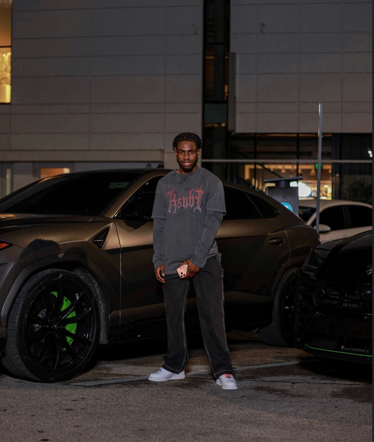 Young man standing in parking lot at night, cars behind him, wearing glasses, hoodie, and sneakers.
