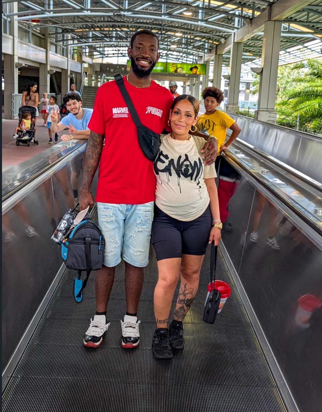 Two people walking on an airport moving walkway, with children and a glass ceiling in the background.