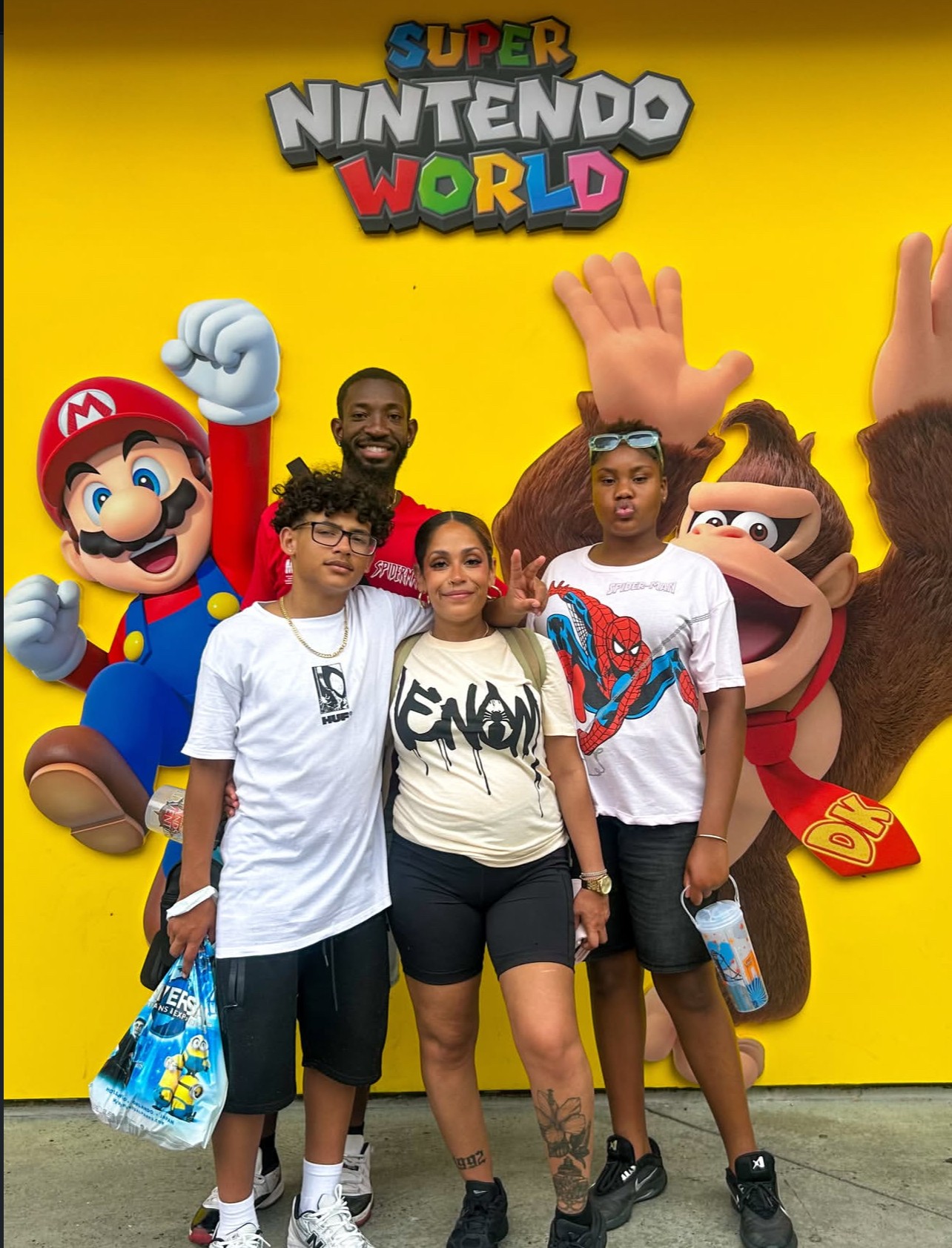 Group of four people standing in front of a Super Nintendo World sign, with colorful characters and yellow background.