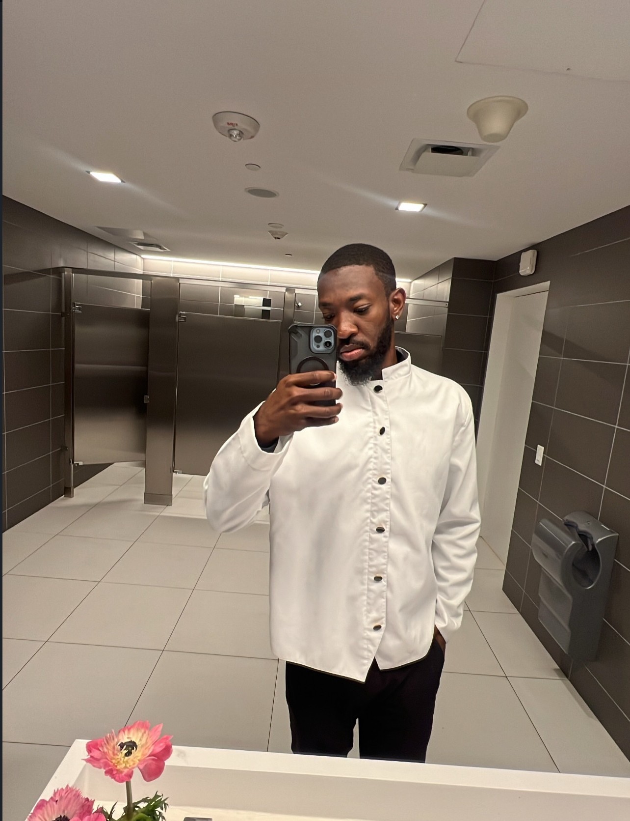 Man taking a selfie in a public restroom with dark tiled walls and a mirror, wearing a white shirt and black pants.
