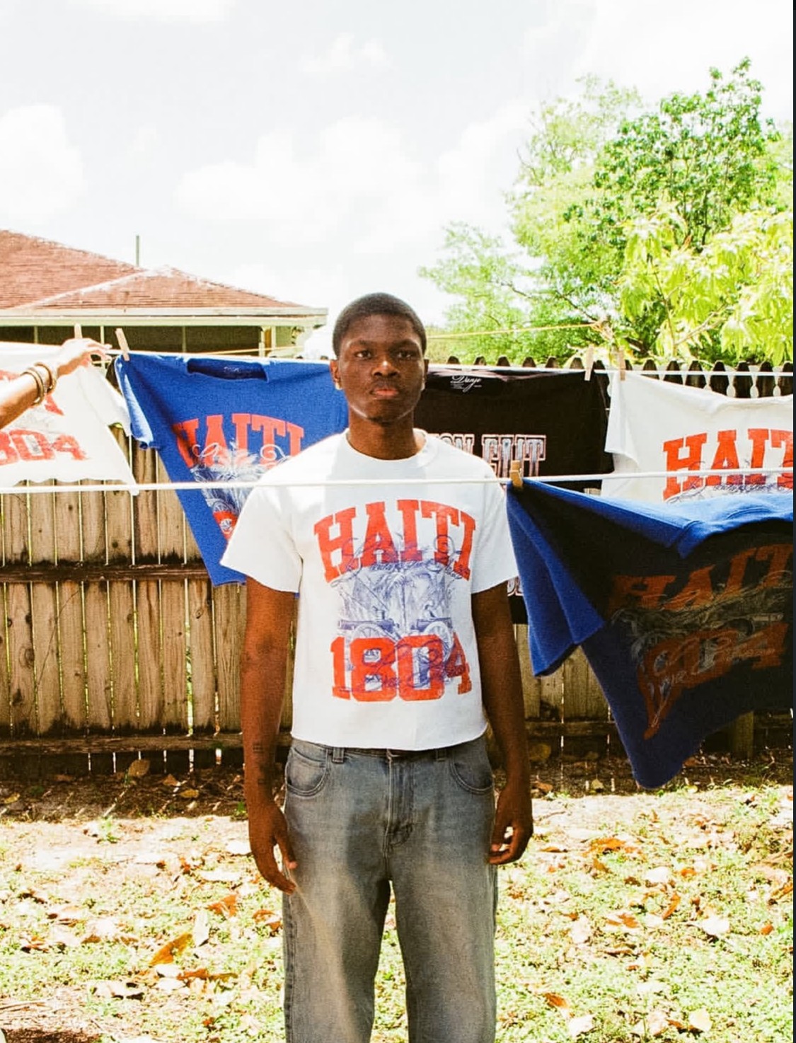 Young man standing outdoors in front of a wooden fence with banners, trees, and a house in the background.