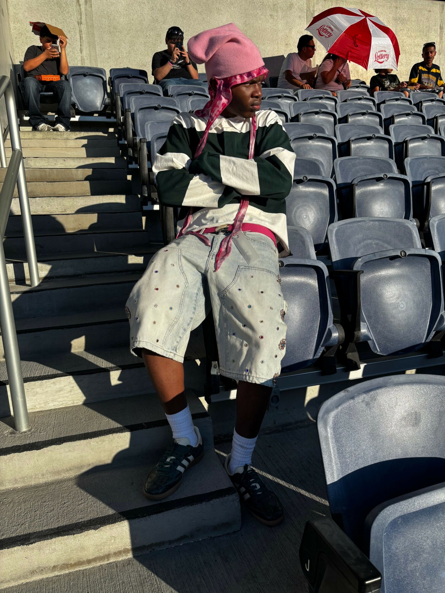 Person wearing a pink headscarf and white coat with black stripes, sitting on stadium seats, holding a pink object.
