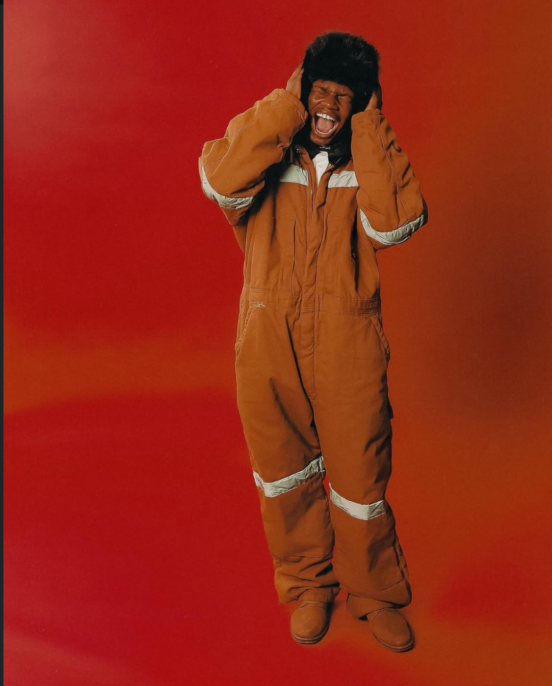 Person in a brown jumpsuit with hands on head, smiling against a red background.
