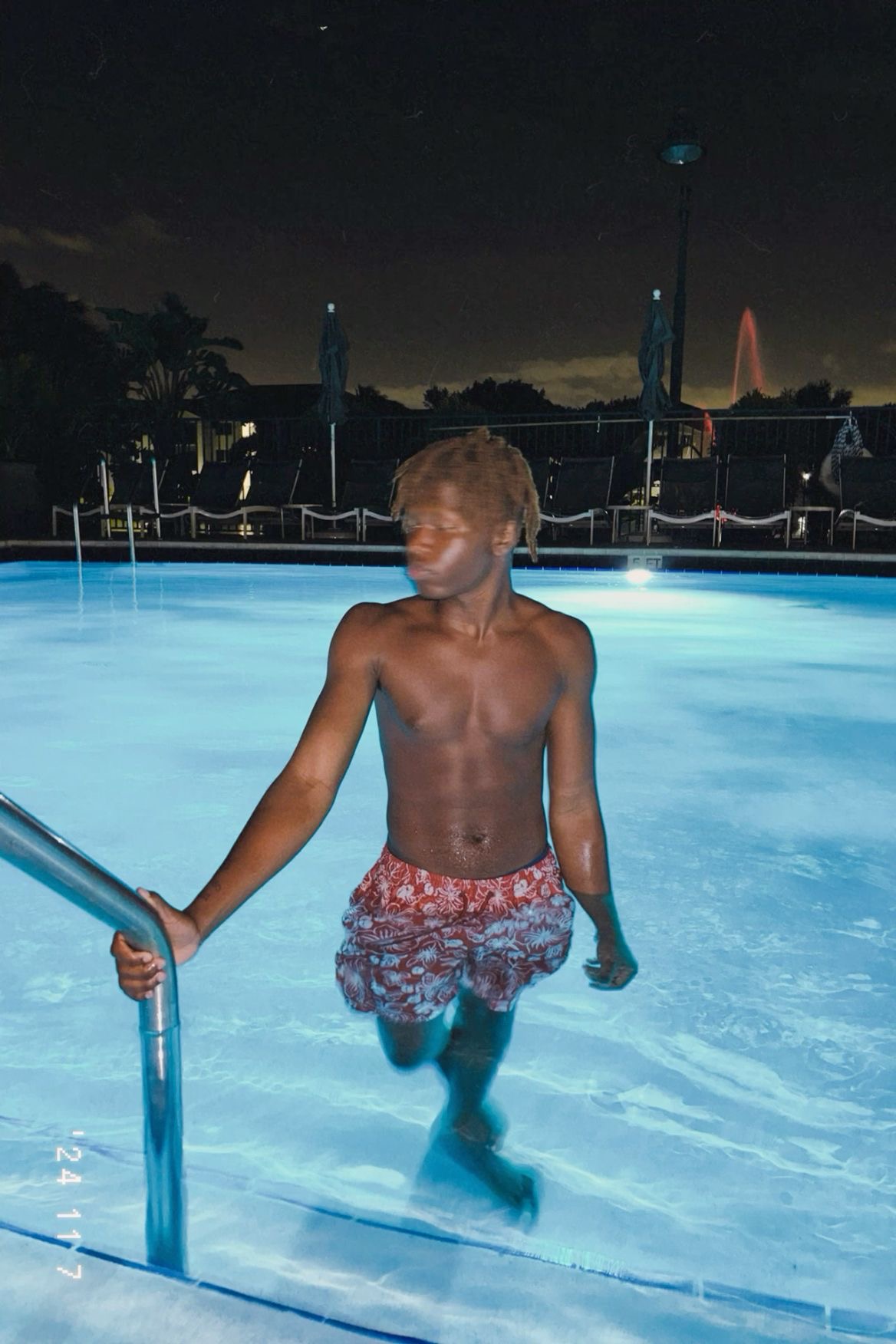 Child walking in a swimming pool at night, holding a handrail, with a dark sky and pool lights visible.