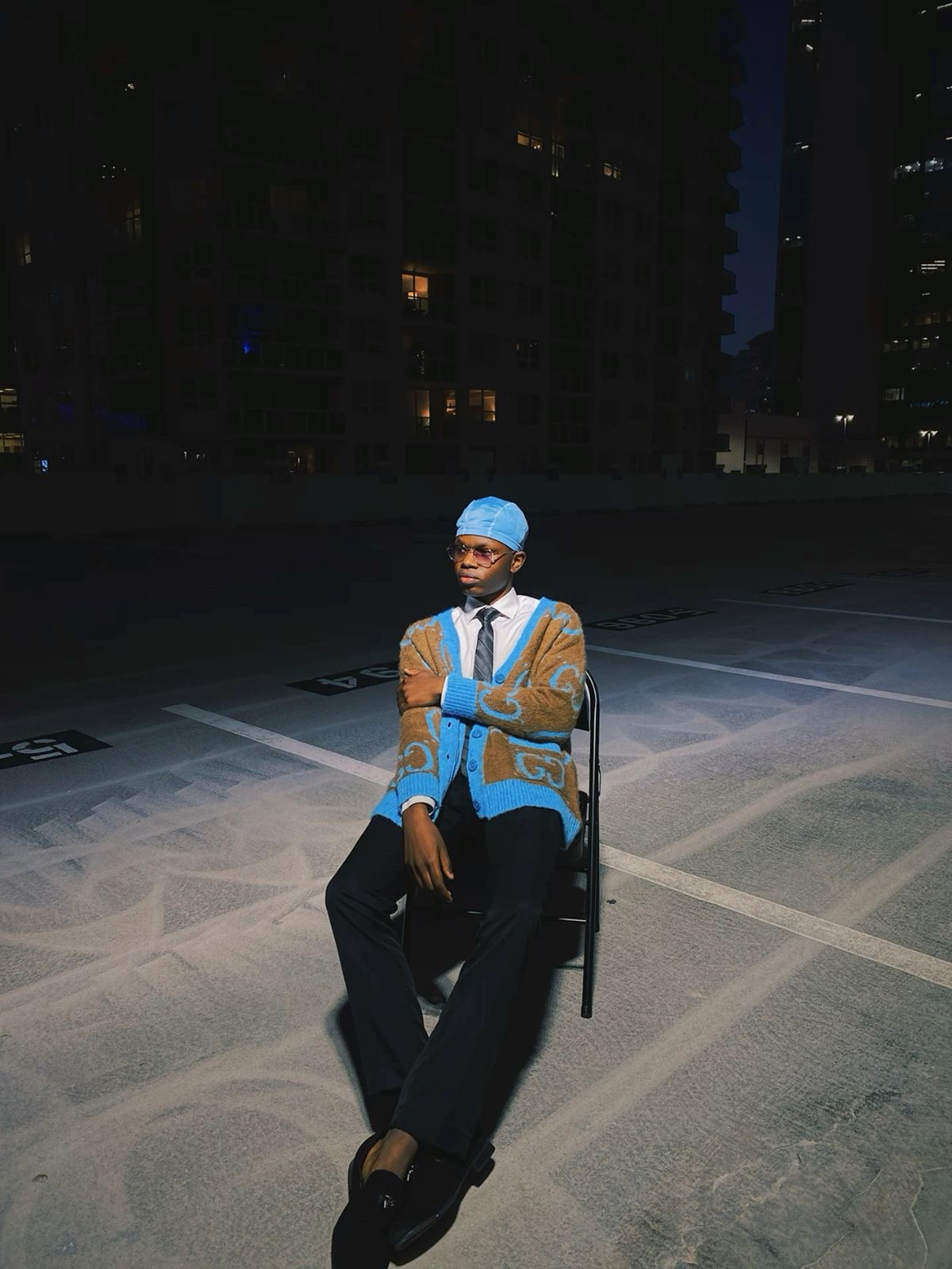 Person sitting on a chair in a parking lot at night, wearing a blue head covering and a patterned coat.