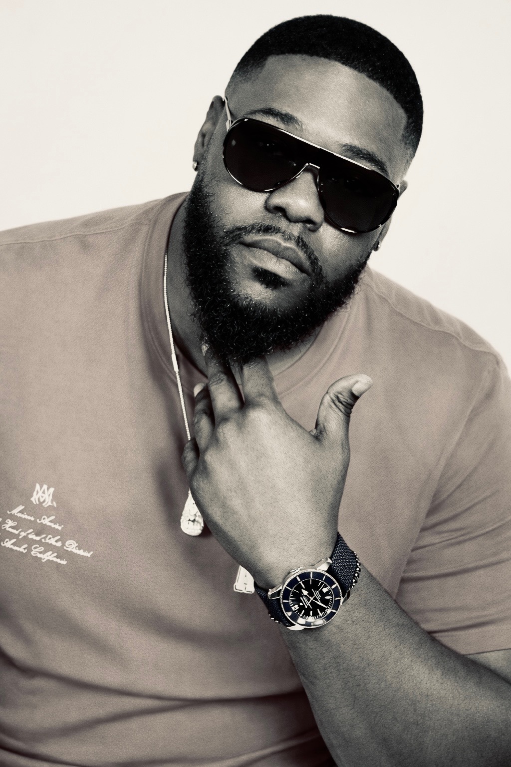 Man with sunglasses, beard, and watch, wearing a t-shirt, posing with hand near chin, against a plain background.