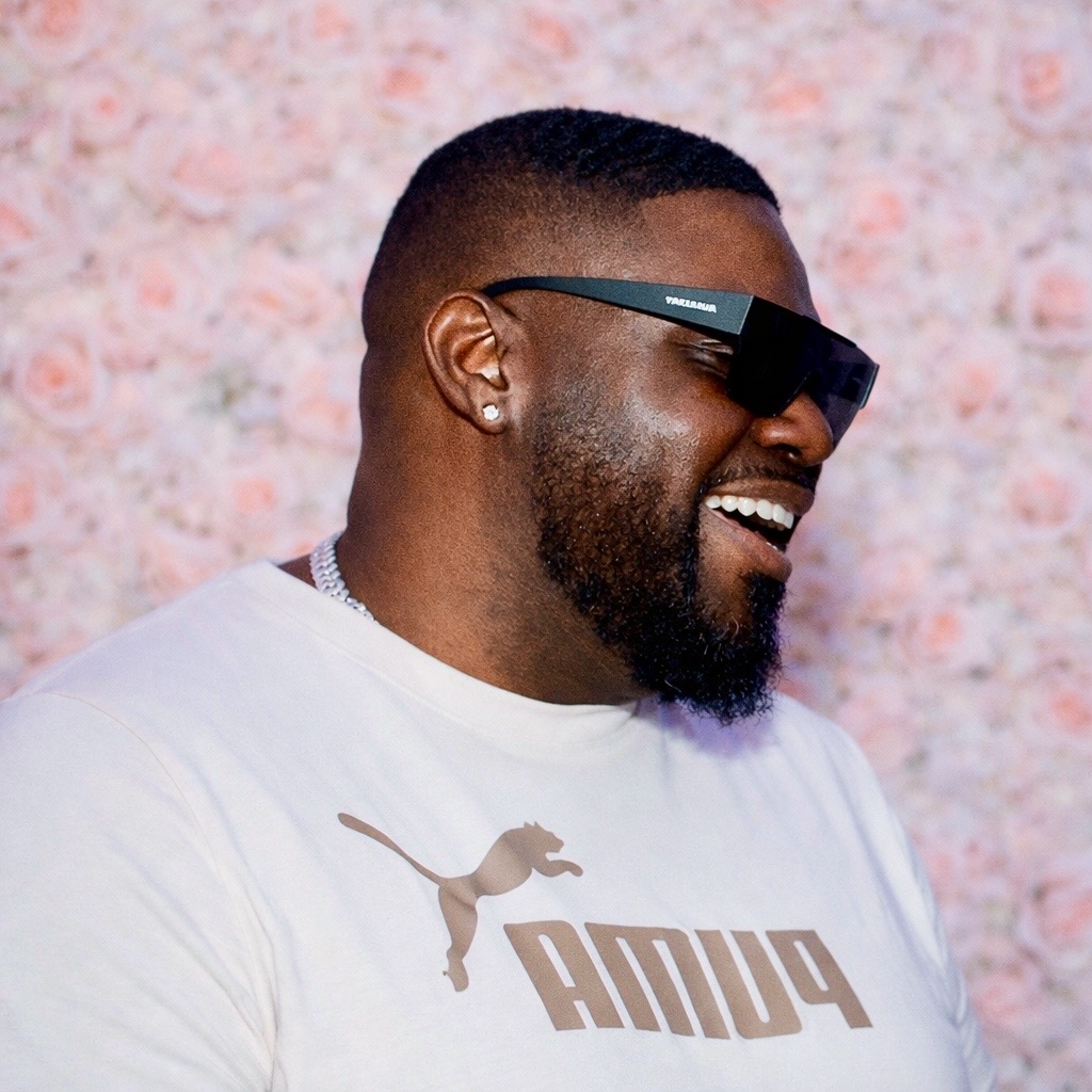 Man with sunglasses smiling, wearing a white Puma shirt, against a pink textured background.