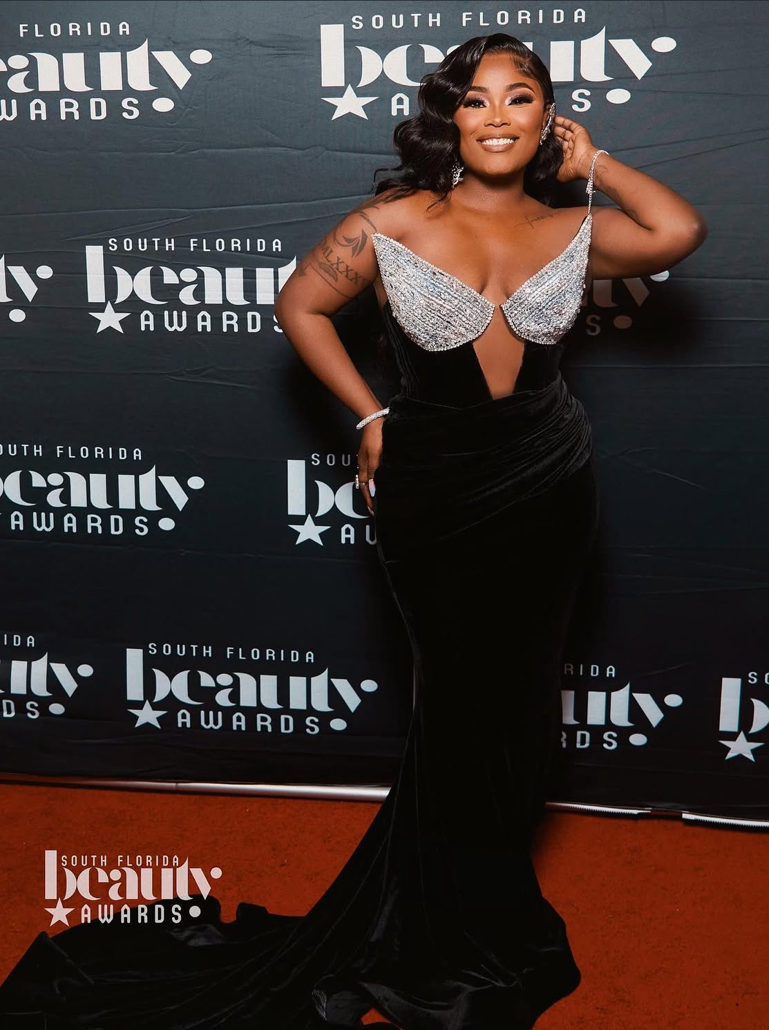 Woman in black gown with silver embellishments on the bodice, smiling at event backdrop, hand on her head.