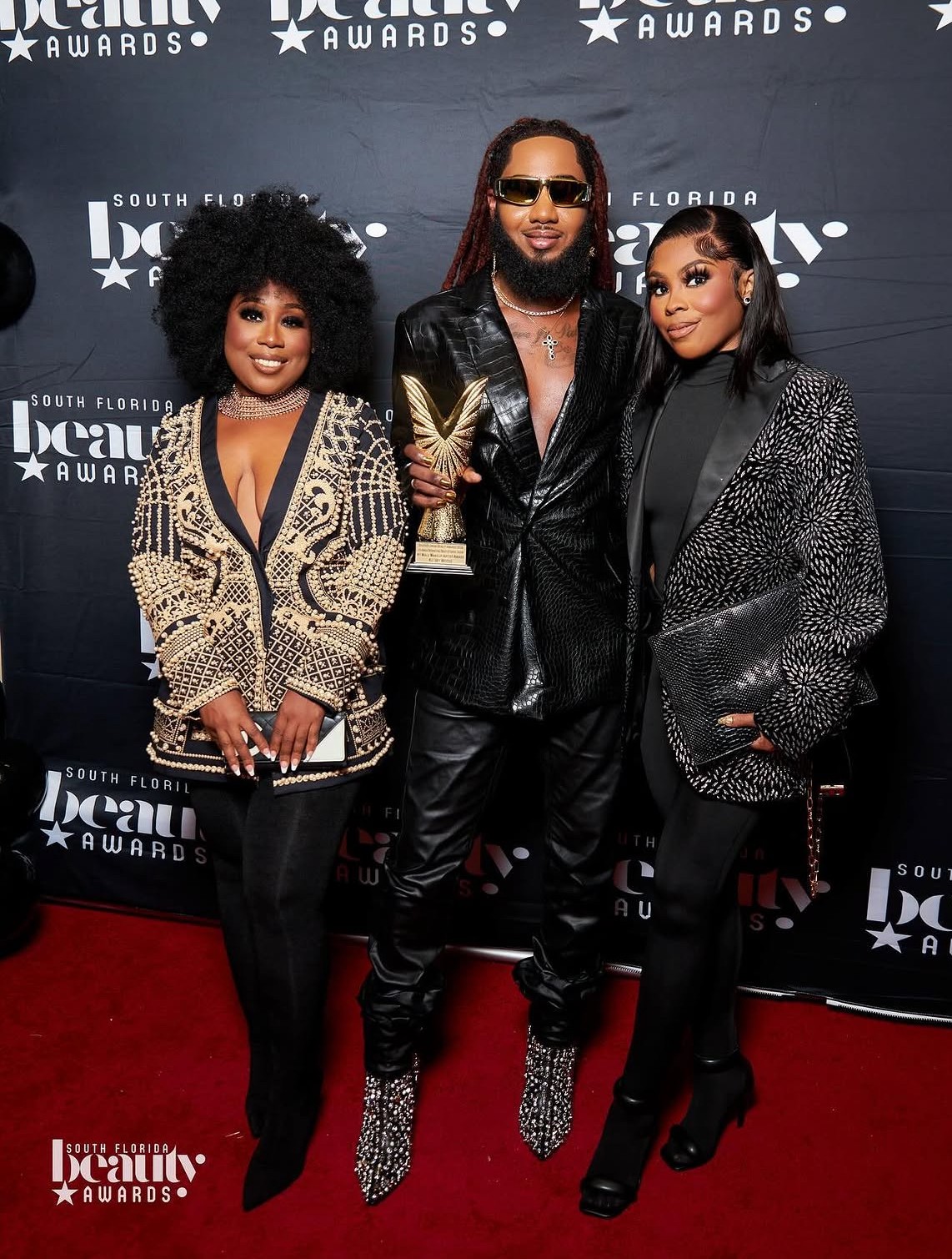 Three people pose on red carpet at awards event, holding a trophy, with a backdrop displaying event logos.