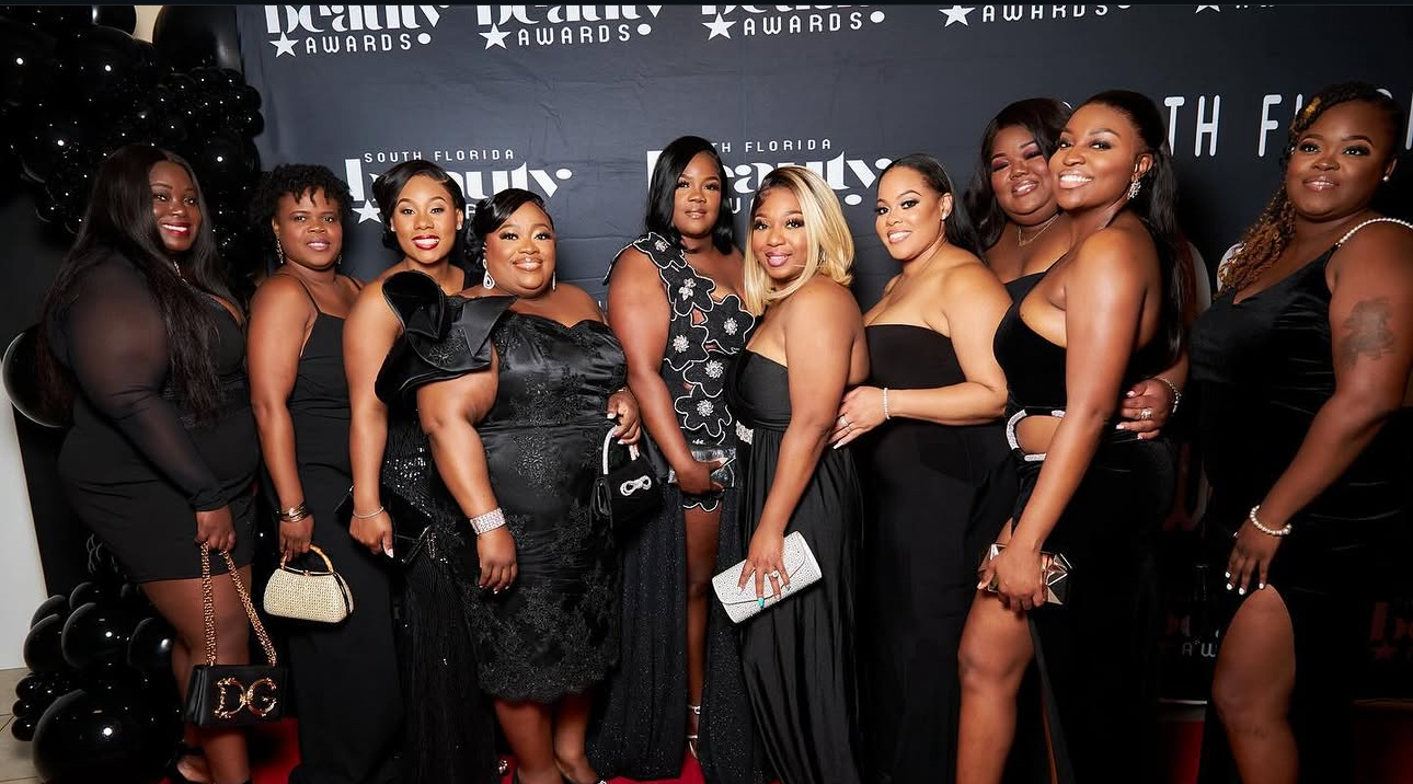 Group of women in black dresses posing at an event with a backdrop, smiling and standing close together.