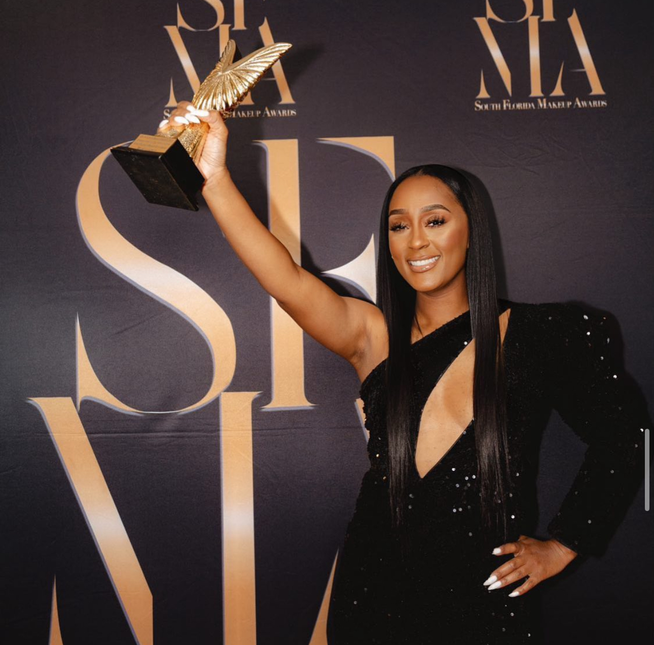 Woman in black dress holding award trophy, smiling, at an event backdrop with text. Word count: 20.