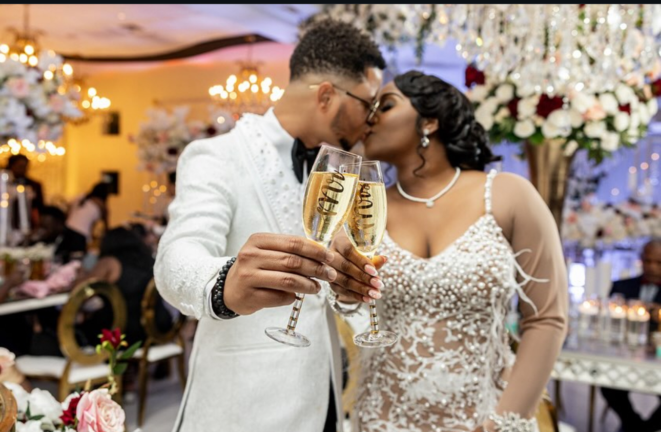 Couple kissing at a celebration, holding champagne glasses, with floral decorations and guests in the background.