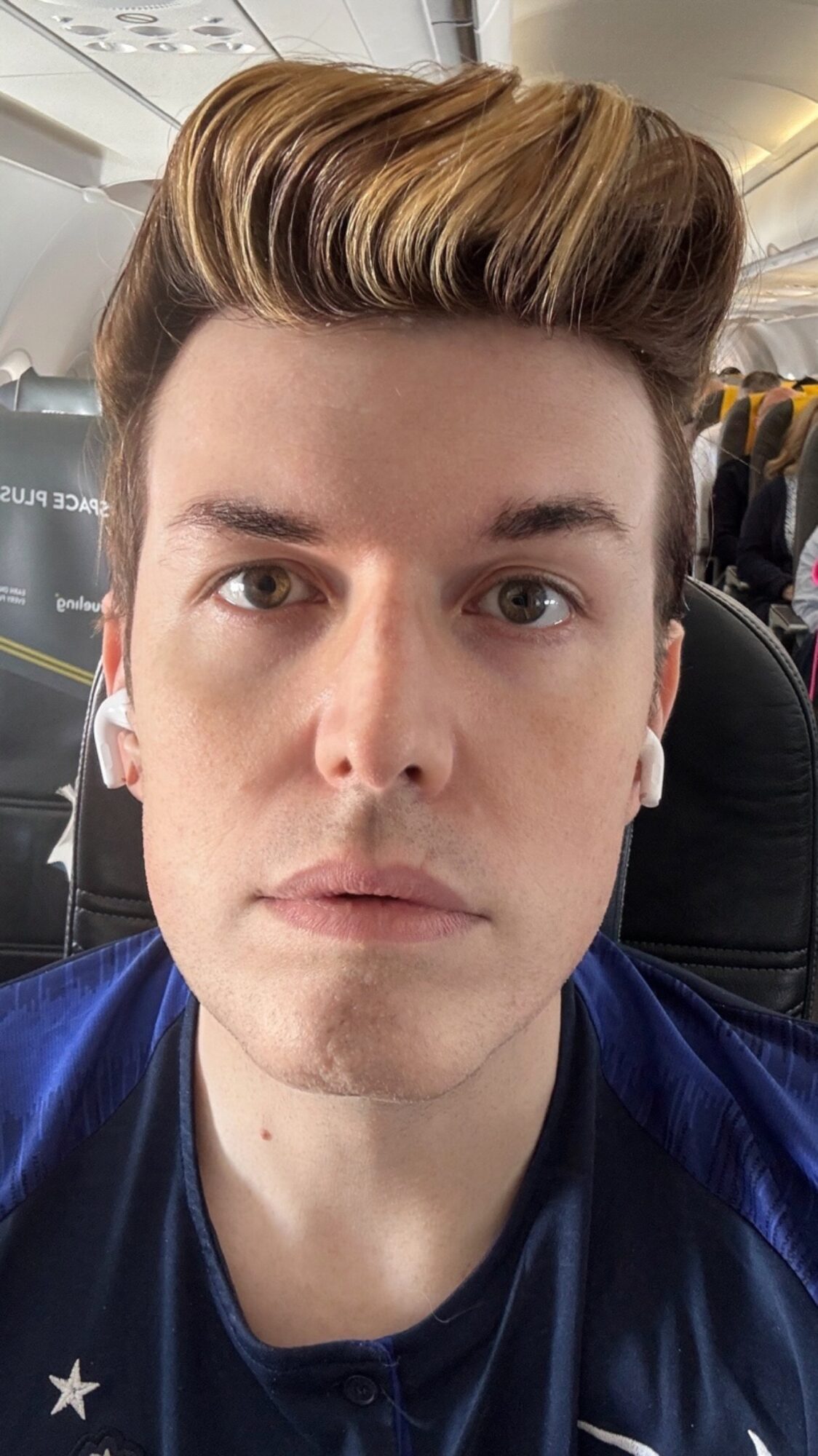 Young man with styled hair, wearing a dark shirt, sitting in an airplane seat, looking directly at the camera.