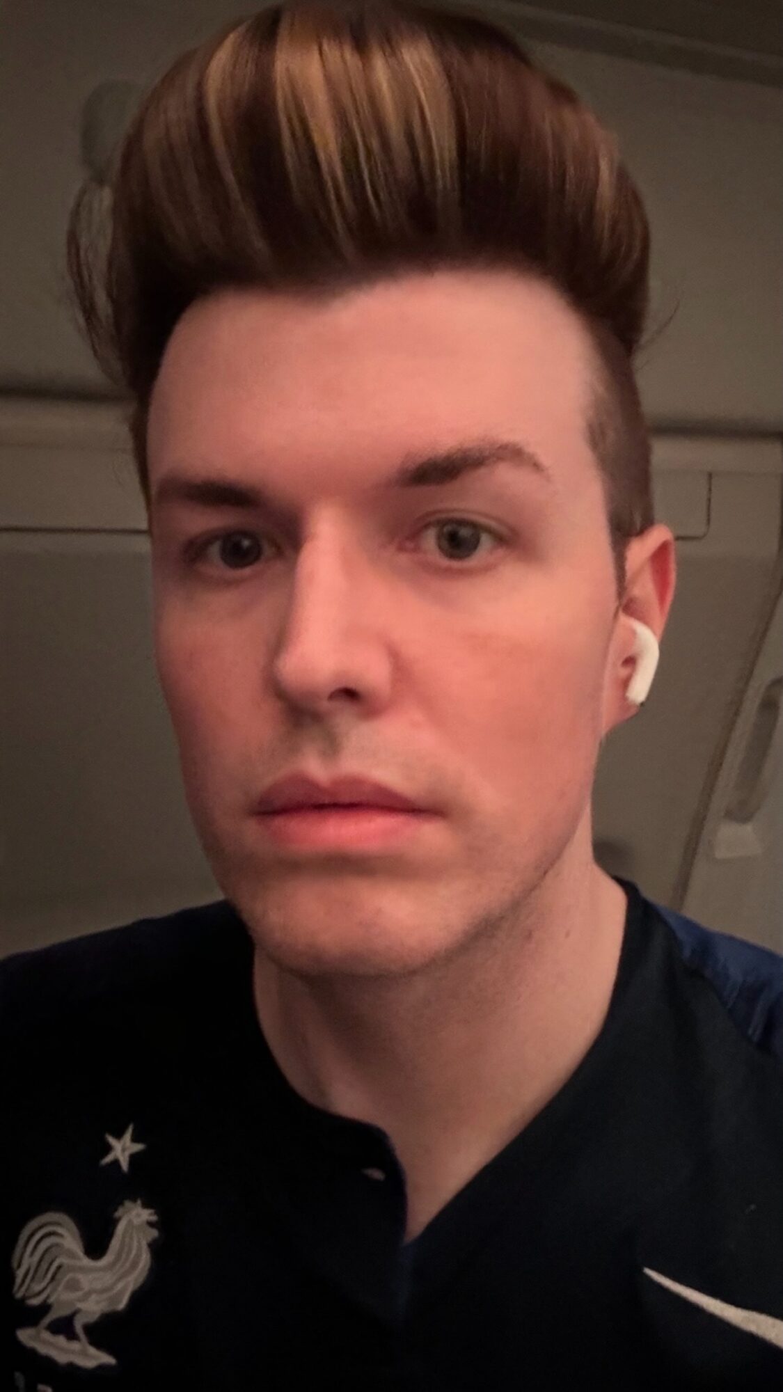 Young man with styled brown hair, wearing a dark shirt, looking directly at the camera, indoors.