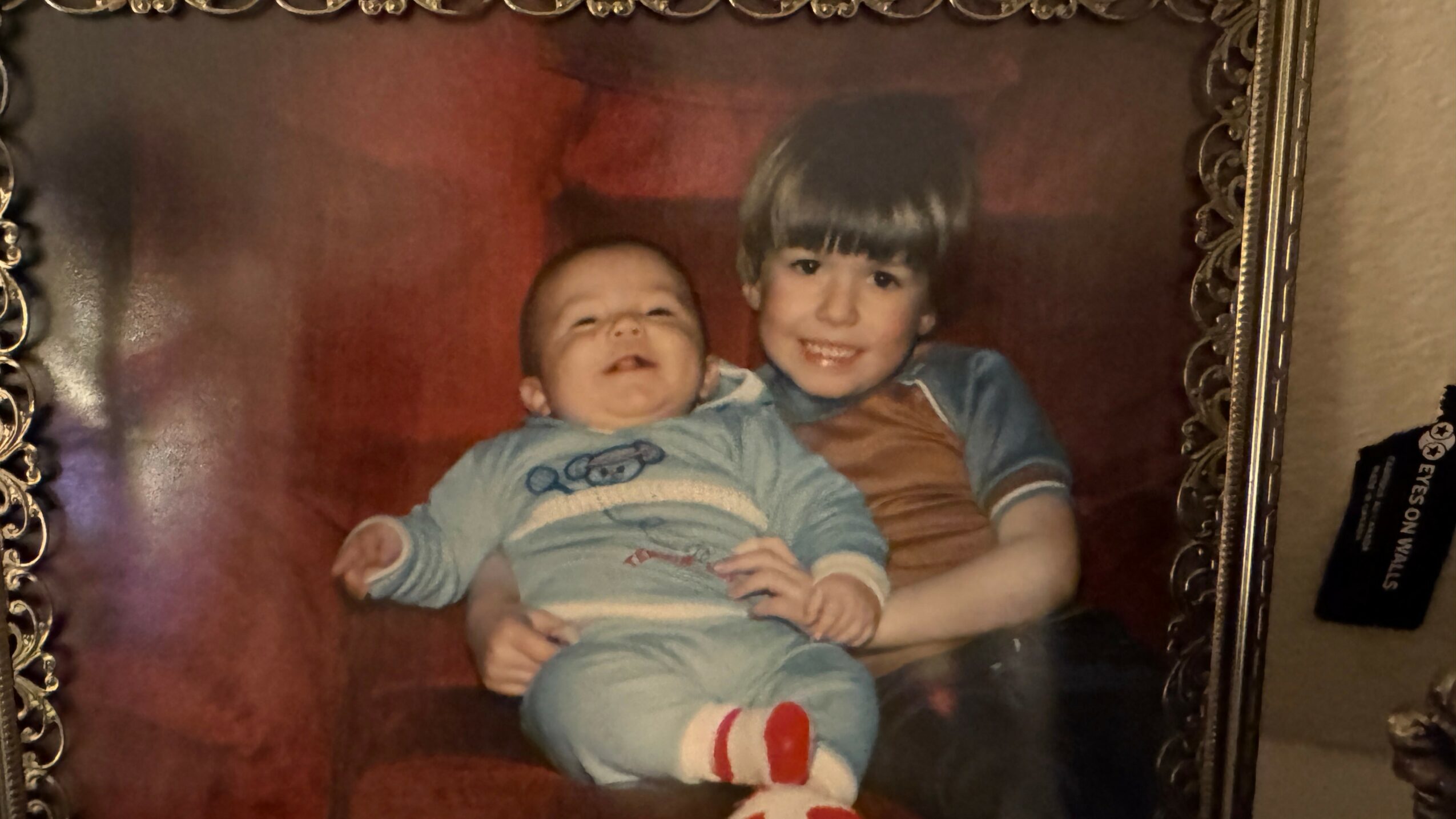 Two children, one older and one younger, sitting together and smiling, in front of a red background.