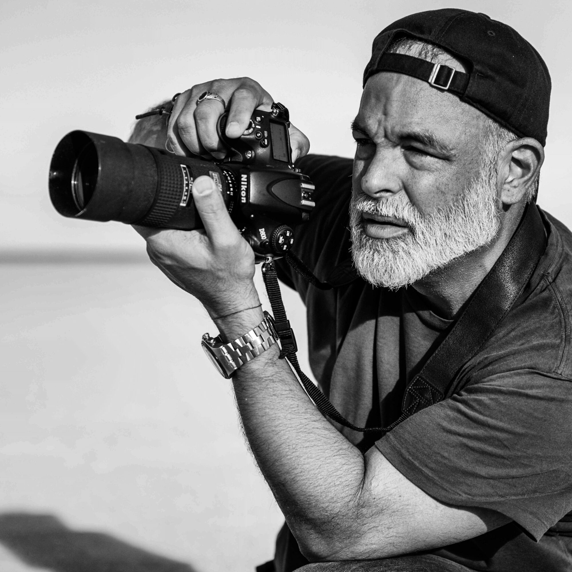 Man with a beard and cap taking a photograph with a camera, outdoors, in black and white.