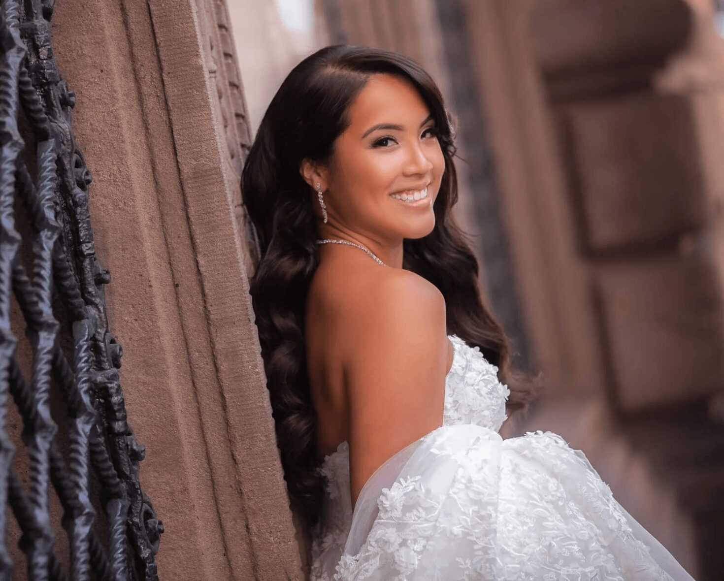 Woman in a white dress smiling, leaning against a stone wall, with dark hair and earrings.
